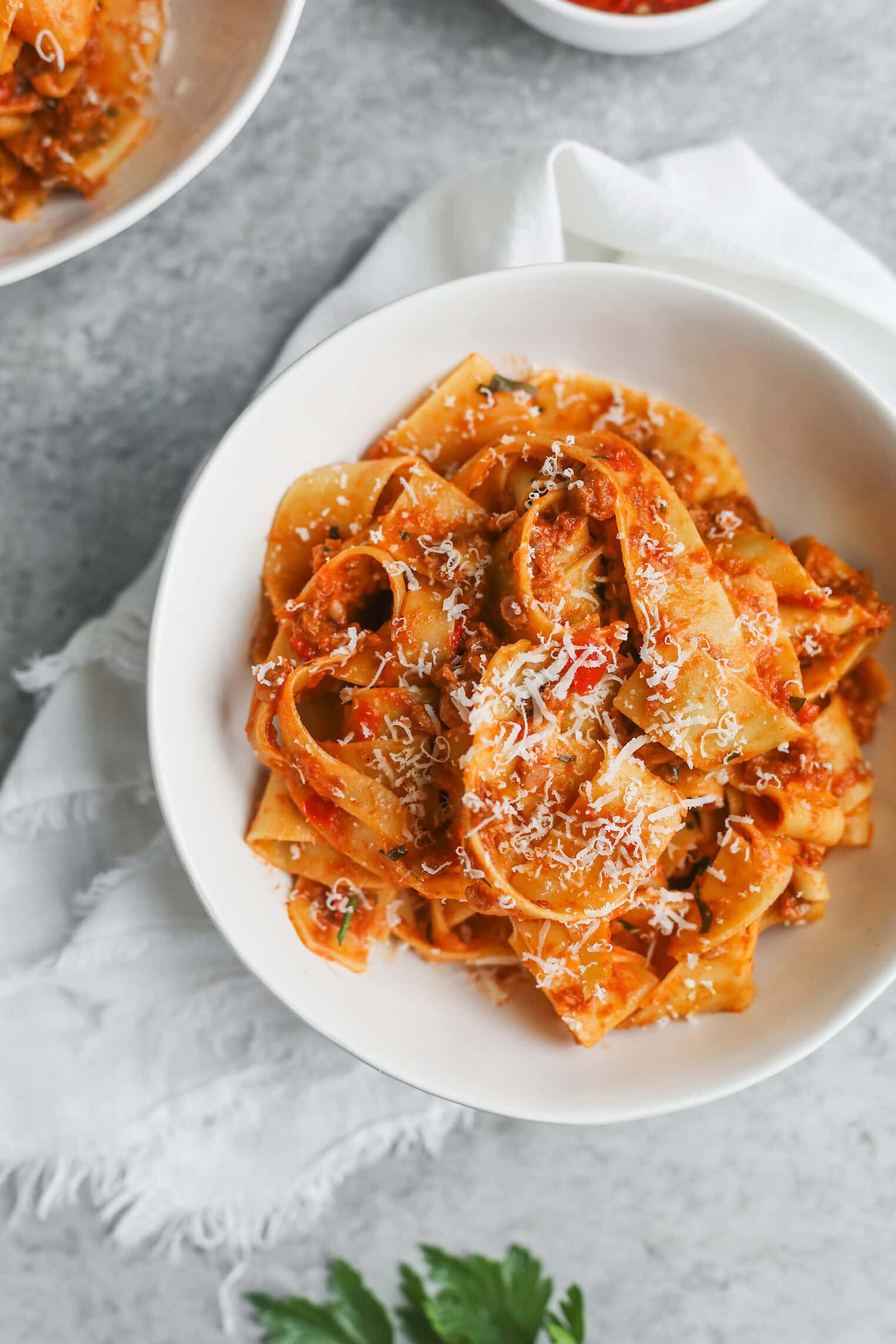 A White Bowl Filled With Wide Pasta Noodles Coated In A Rich Plant-Based Bolognese Sauce, Topped With Grated Cheese, Sits On A White Napkin On A Gray Surface.