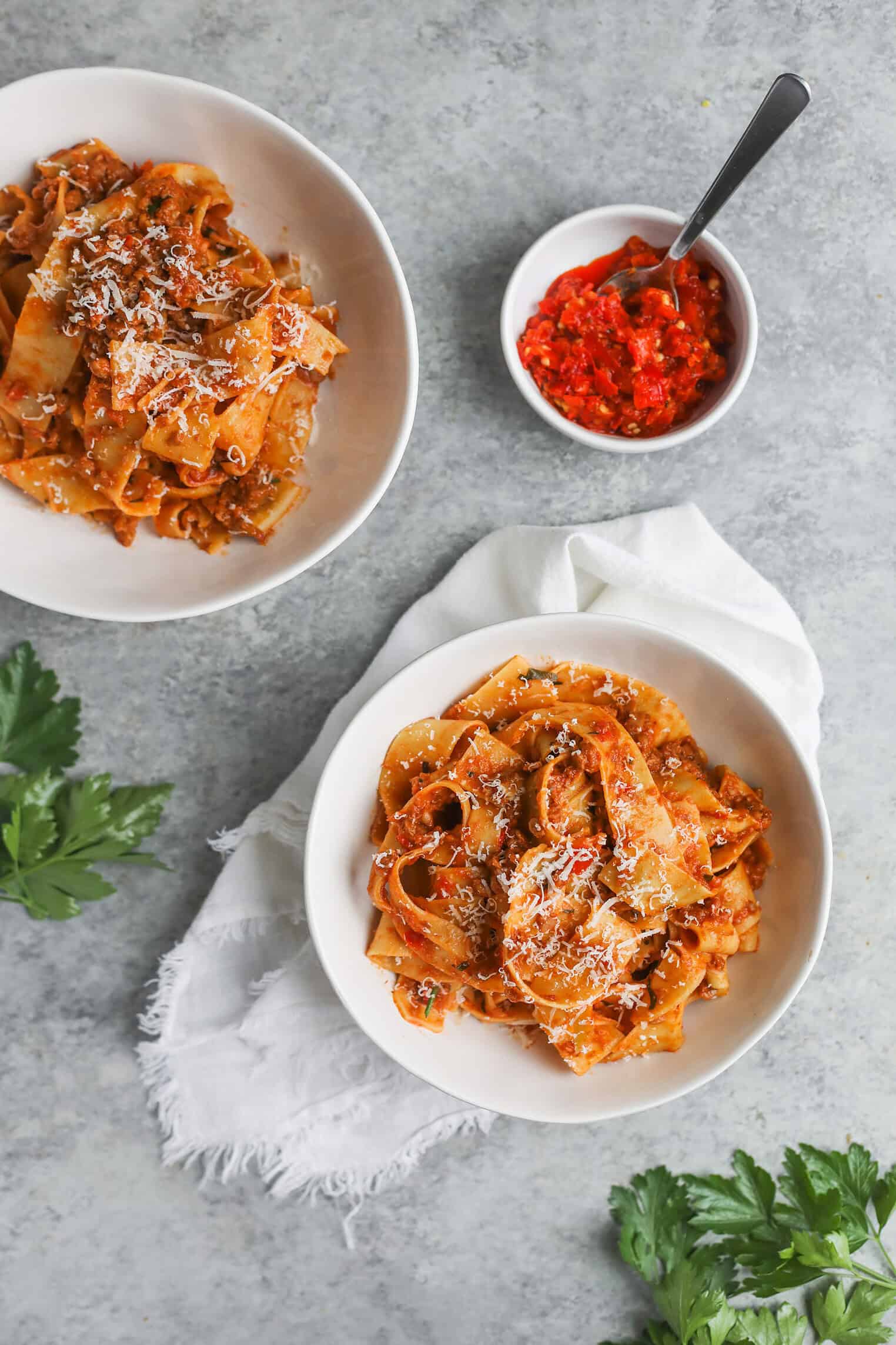 Two White Bowls Filled With Wide, Ribbon-Like Pasta Topped With Vegan Bolognese Sauce And Grated Cheese. A Small Bowl Of Red Chili Paste With A Spoon Sits Nearby On A Light Surface, With Green Parsley Leaves As Garnish.