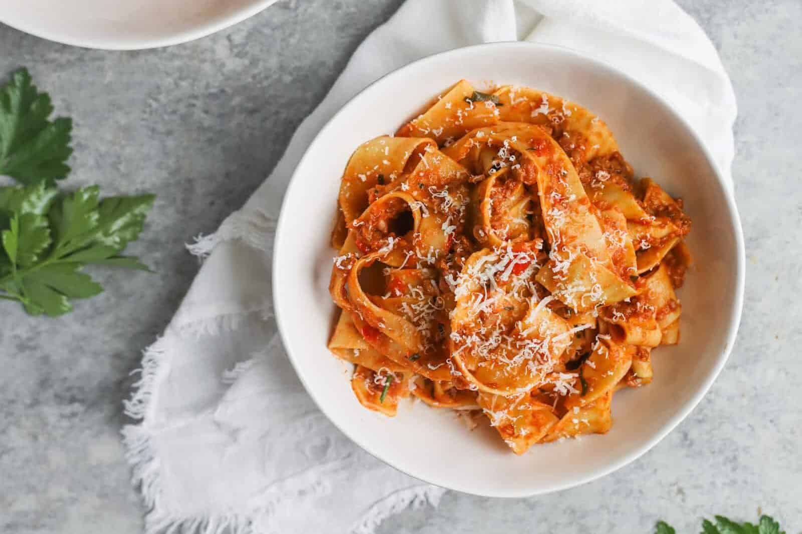 Two White Bowls Filled With Wide, Ribbon-Like Pasta Topped With Vegan Bolognese Sauce And Grated Cheese. A Small Bowl Of Red Chili Paste With A Spoon Sits Nearby On A Light Surface, With Green Parsley Leaves As Garnish.