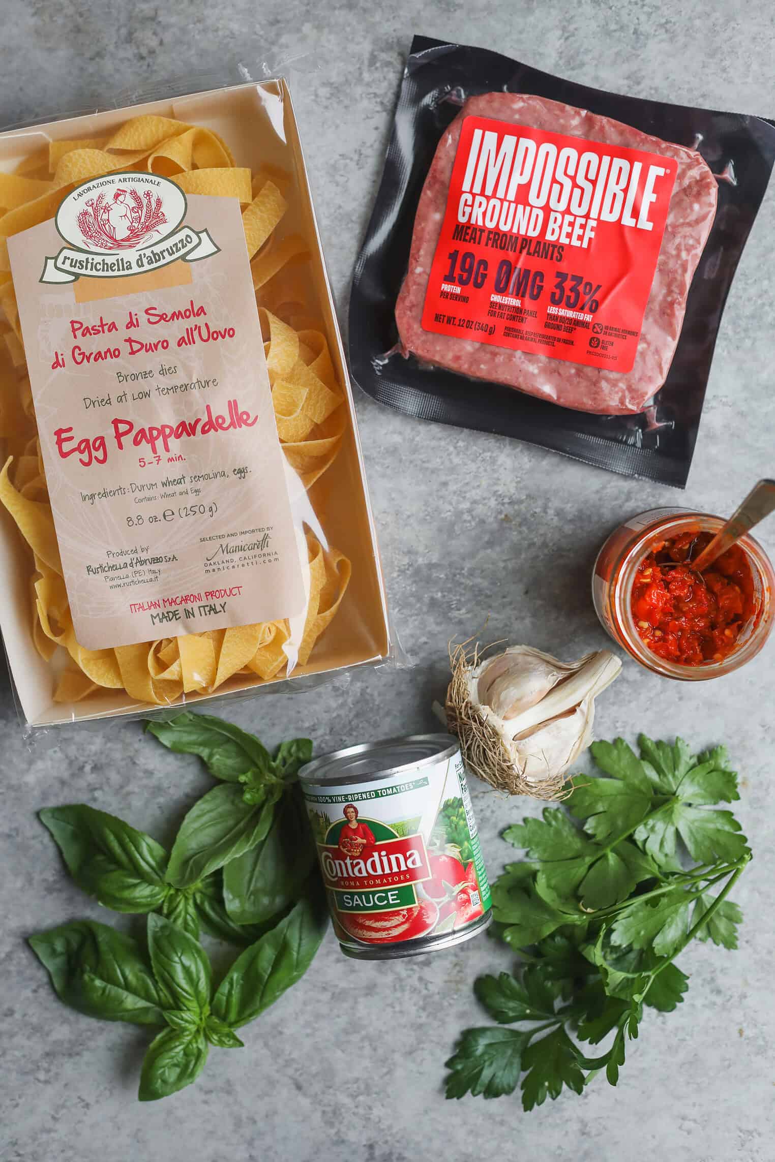 A Flat Lay Of Pasta Ingredients: Egg Pappardelle, Impossible Ground Beef For A Delicious Plant-Based Bolognese, A Jar Of Red Sauce, Tomato Sauce, Fresh Basil, Parsley, And Garlic On A Gray Surface.