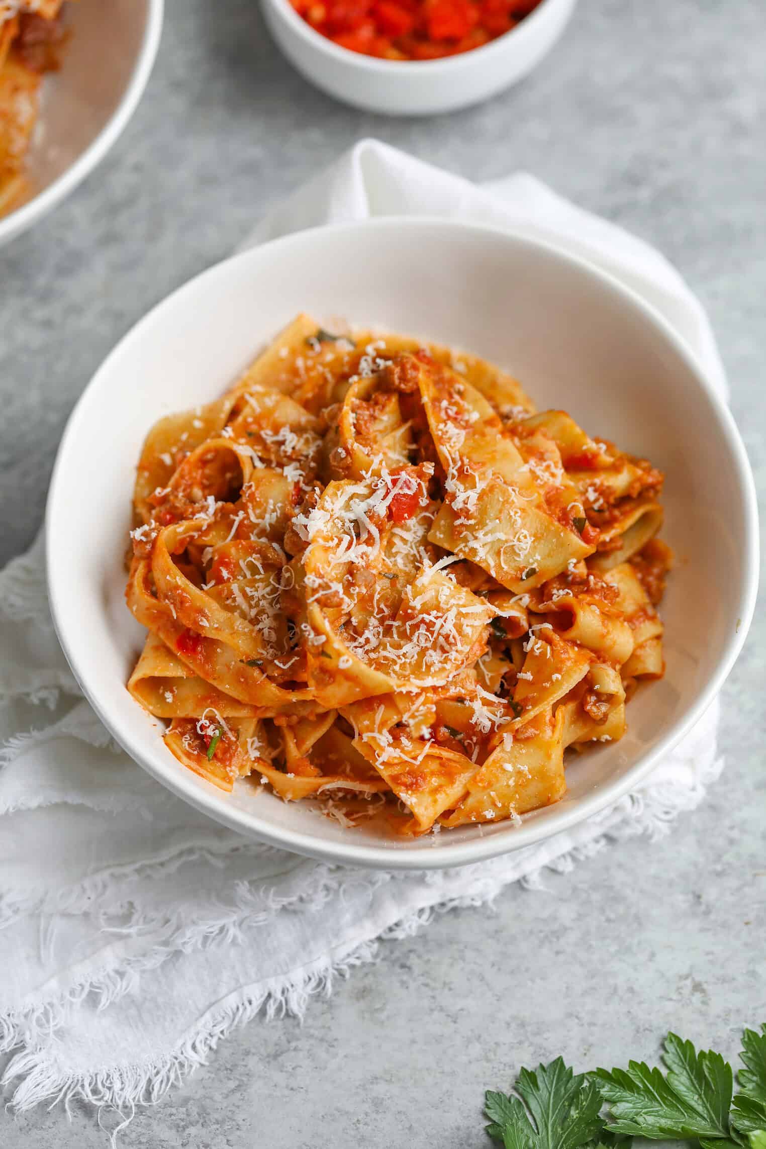 A White Bowl Filled With Wide Ribbon Vegan Pasta Coated In Red Tomato Sauce And Topped With Grated Cheese, Placed On A Textured White Napkin With A Sprig Of Parsley Nearby.