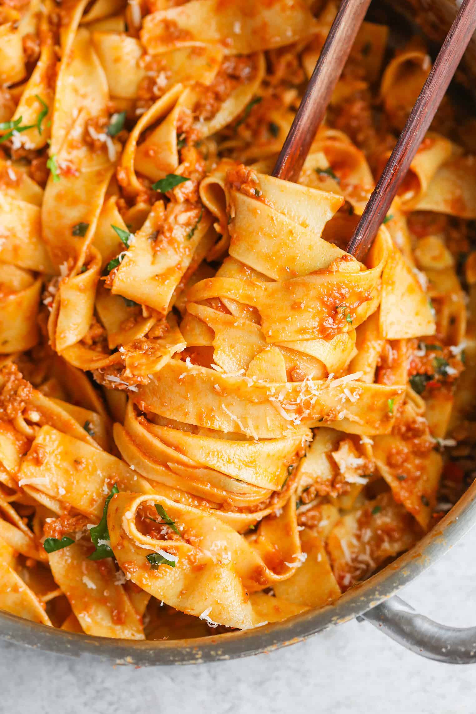 Wide Ribbons Of Pappardelle Pasta Coated In A Rich, Chunky Impossible Bolognese Sauce, Garnished With Grated Cheese And Parsley, Being Twirled With Wooden Tongs Inside A Metal Pan.