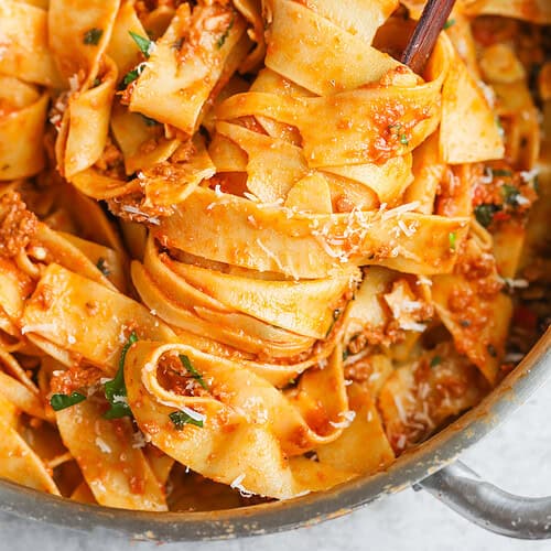 Wide Ribbons Of Pappardelle Pasta Coated In A Rich, Chunky Impossible Bolognese Sauce, Garnished With Grated Cheese And Parsley, Being Twirled With Wooden Tongs Inside A Metal Pan.