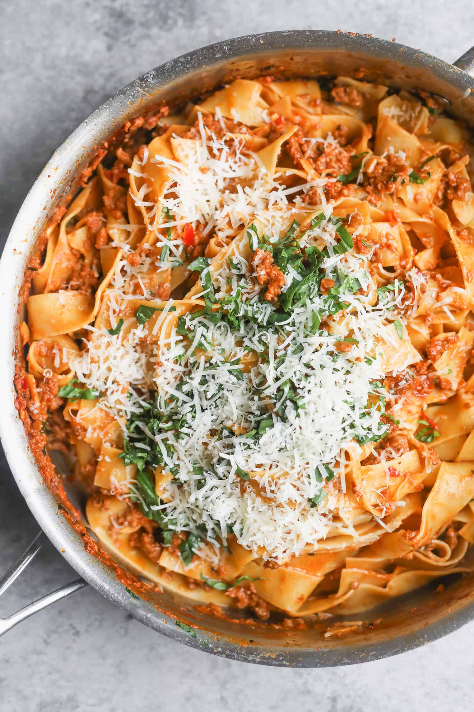 A Skillet Filled With Wide Pasta Noodles In A Creamy Plant-Based Bolognese Sauce, Topped With Shredded Parmesan Cheese And Chopped Fresh Basil.