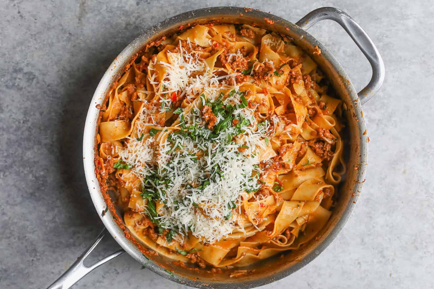 A Stainless Steel Pan Filled With Pappardelle Vegan Pasta In A Rich Red Impossible Bolognese Sauce, Topped With Grated Parmesan Cheese And Chopped Herbs, Sits On A Gray Countertop.