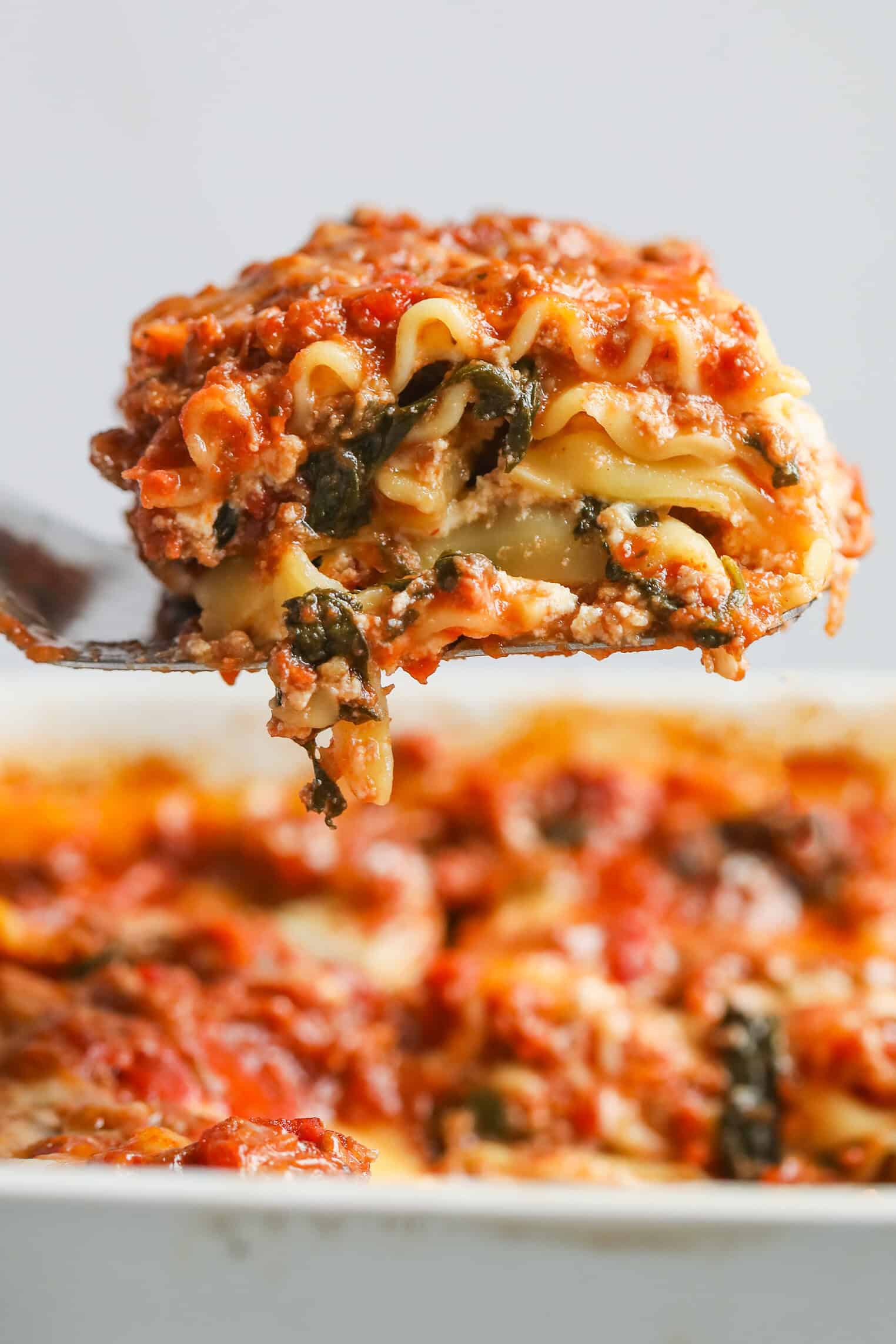 Impossible Lasagna 1 A Close-Up Of A Spatula Holding A Slice Of Cheesy Impossible Lasagna Above A Baking Dish Filled With More Lasagna, Showcasing Layers Of Pasta, Spinach, Tomato Sauce, And Melted Cheese.