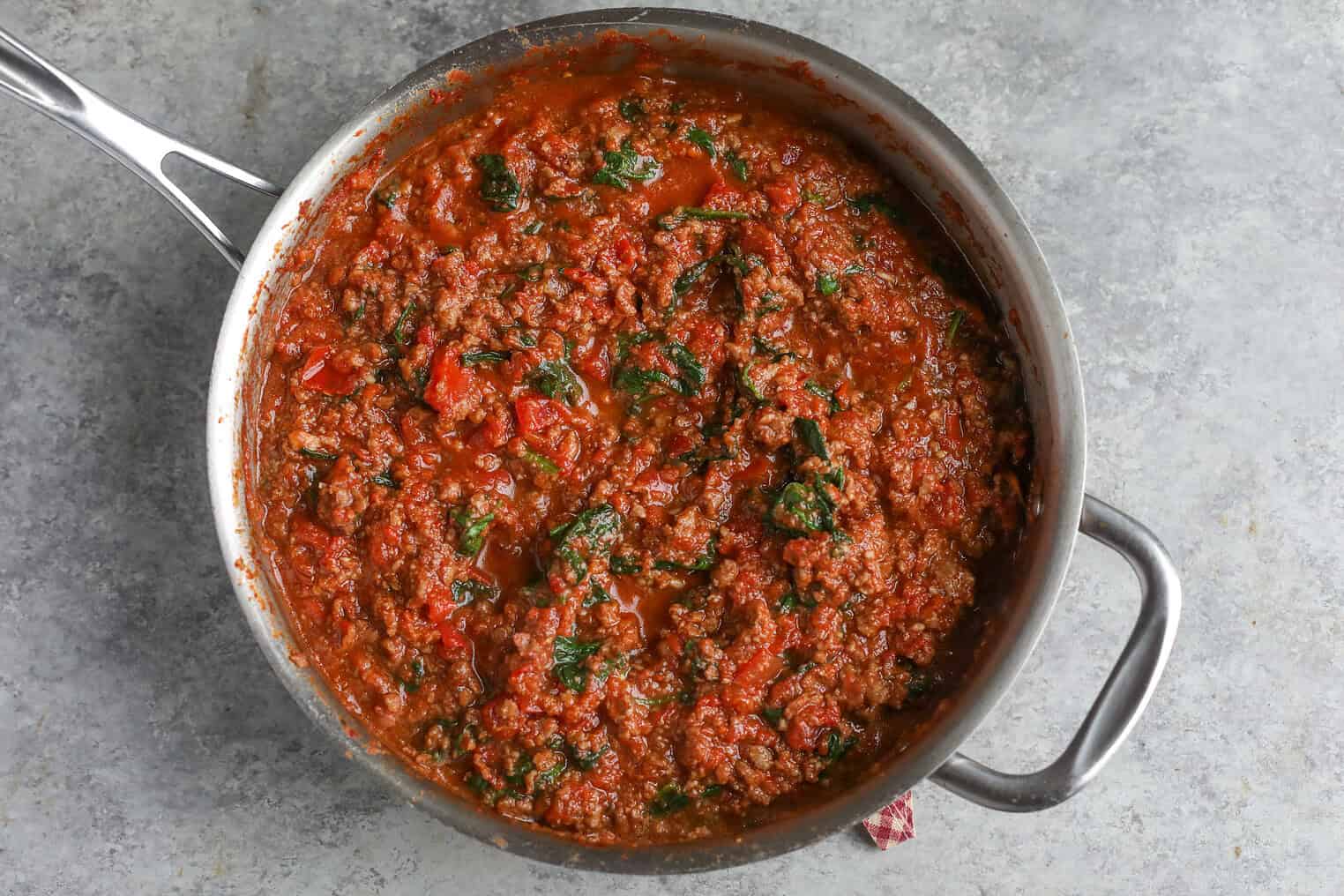 Impossible Lasagna 9 A Stainless Steel Pan Filled With Chunky Tomato Meat Sauce For Impossible Lasagna, Featuring Ground Beef, Tomatoes, And Spinach, Sits On A Gray Textured Surface.
