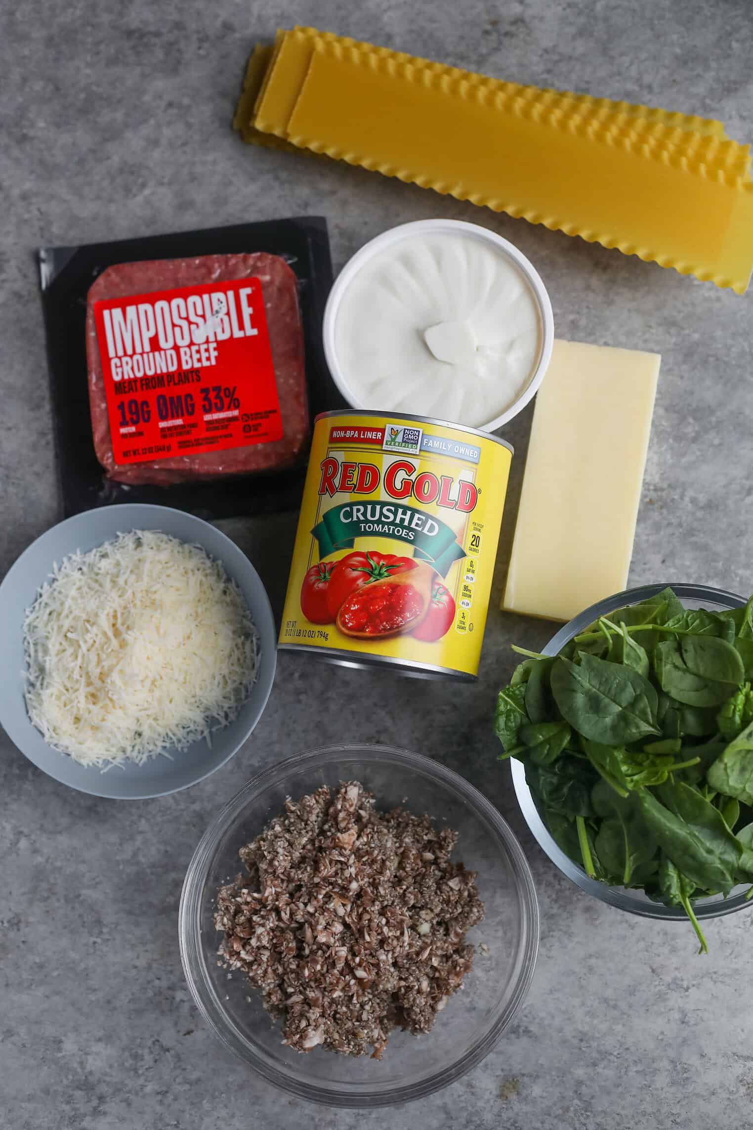 Impossible Lasagna 2 A Top-Down View Of Impossible Lasagna Ingredients: Lasagna Noodles, Impossible Ground Beef, Sour Cream, A Block Of Cheese, Crushed Tomatoes, Shredded Cheese, Chopped Mushrooms, And Fresh Spinach On A Gray Surface.