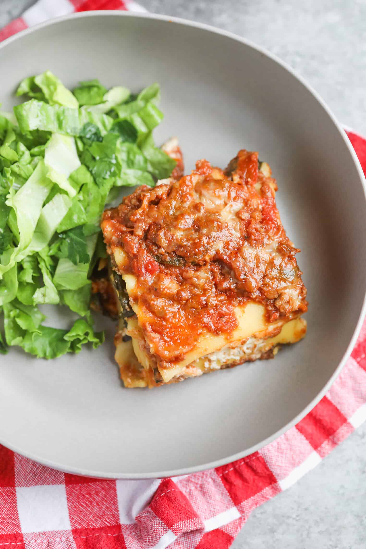 Impossible Lasagna 17 A Plate With A Serving Of Impossible Lasagna Topped With Tomato Sauce Sits Next To Chopped Green Lettuce, All Placed On A Gray Surface With A Red And White Checkered Cloth Underneath.