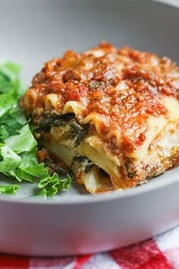 A Close-Up Of Impossible Lasagna Featuring Layers Of Pasta, Plant-Based Meat Sauce, Cheese, And Spinach, Served With A Side Of Fresh Green Lettuce On A Gray Plate.