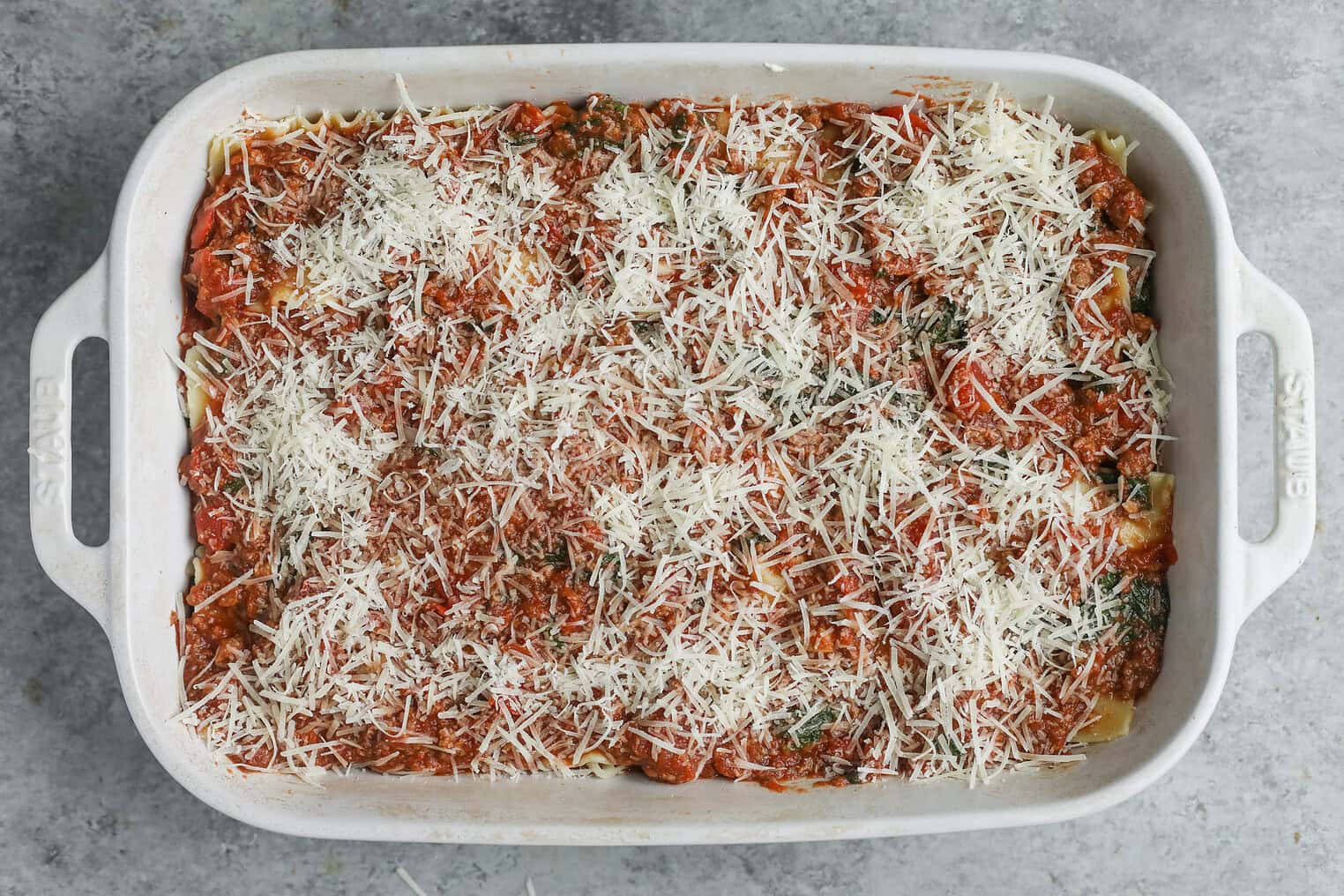 Impossible Lasagna 13 A Rectangular White Baking Dish Filled With Unbaked Impossible Lasagna, Topped With Tomato Sauce, Herbs, And A Generous Layer Of Shredded Cheese, Sits On A Gray Textured Surface.