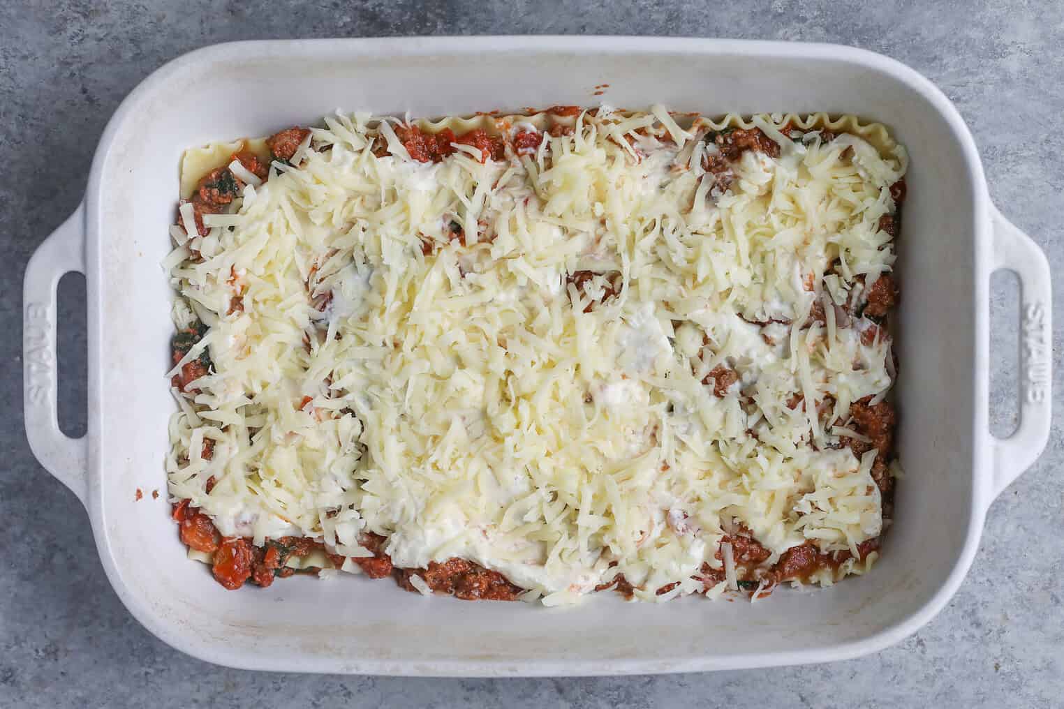 Impossible Lasagna 12 A White Rectangular Baking Dish Filled With Unbaked Impossible Lasagna, Topped With Shredded Cheese And Sauce, Sits On A Gray Countertop.