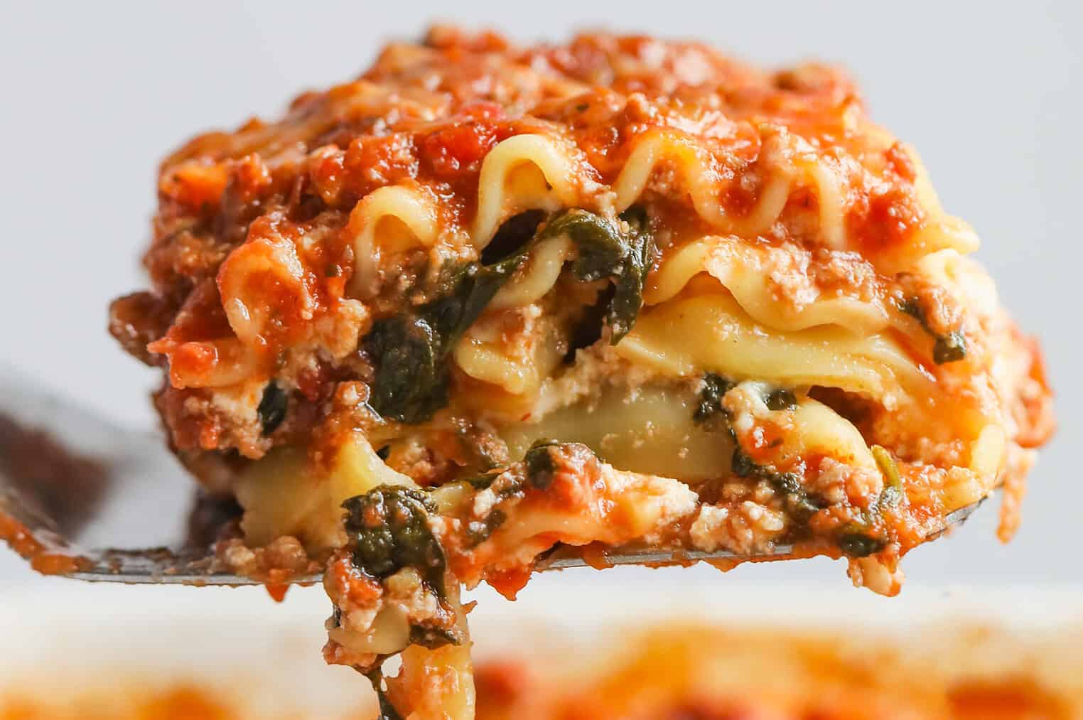 A Close-Up Of A Fork Lifting A Cheesy, Layered Slice Of Impossible Lasagna Filled With Ricotta, Spinach, Meat Sauce, And Pasta, With Sauce And Melted Cheese Visible.