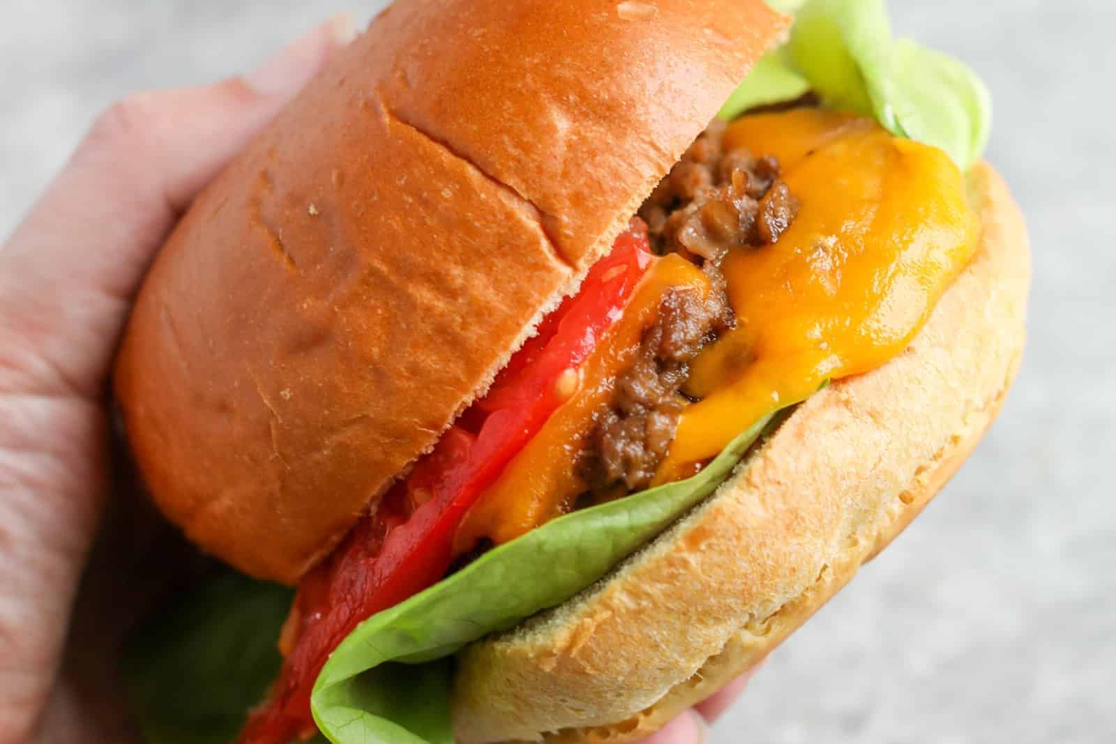A Hand Holding An Impossible Smash Burger With Lettuce, Tomato, Ground Beef, And Melted Cheddar Cheese In A Soft Bun.