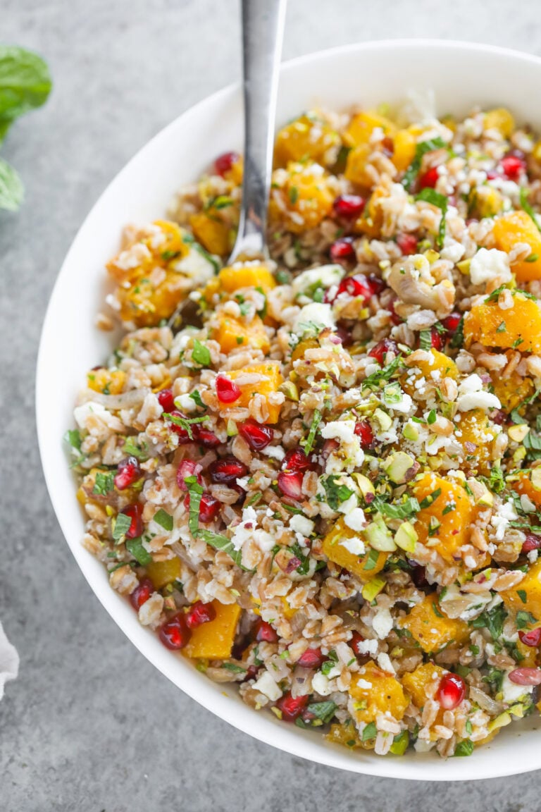 A Bowl Of Jeweled Farro Salad With Roasted Butternut Squash, Pomegranate Seeds, Chopped Herbs, Crumbled Cheese, And A Fork Resting Inside The Bowl.