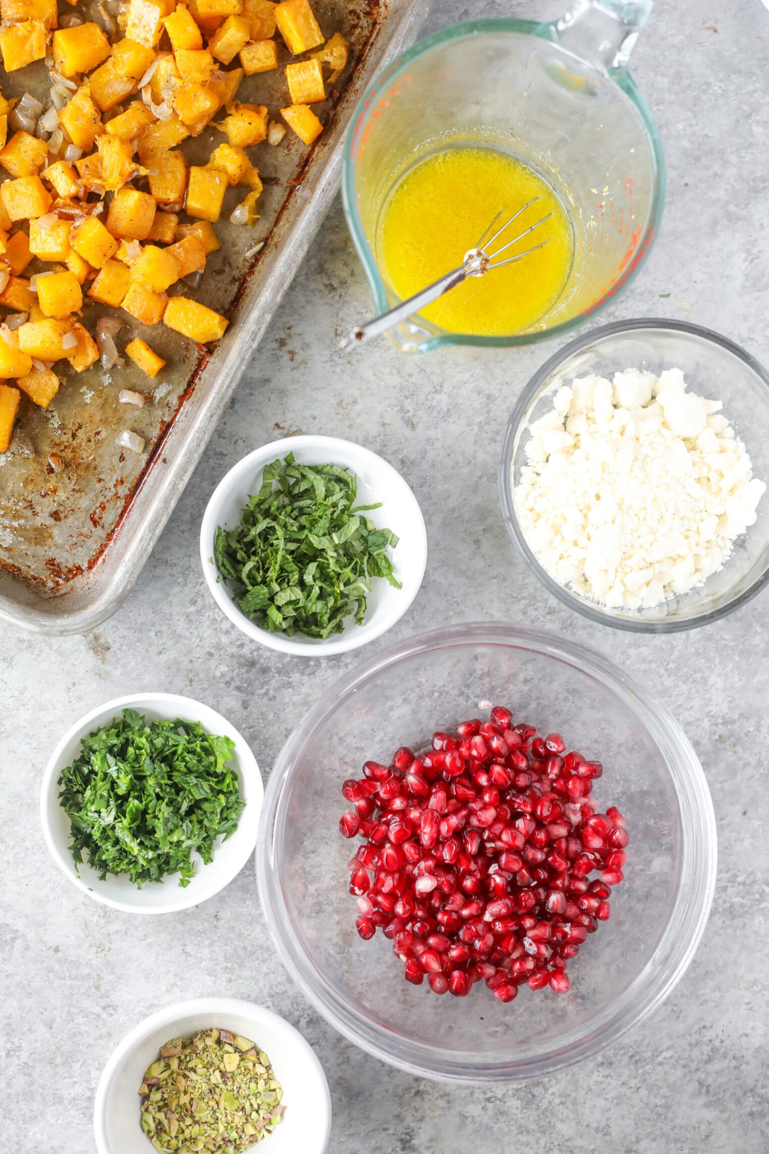 Overhead View Of Ingredients For A Jeweled Farro Salad On A Gray Surface: Roasted Butternut Squash On A Tray, A Measuring Cup With Whisked Dressing, Bowls Of Feta Cheese, Pomegranate Seeds, Chopped Herbs, And Shelled Pistachios.