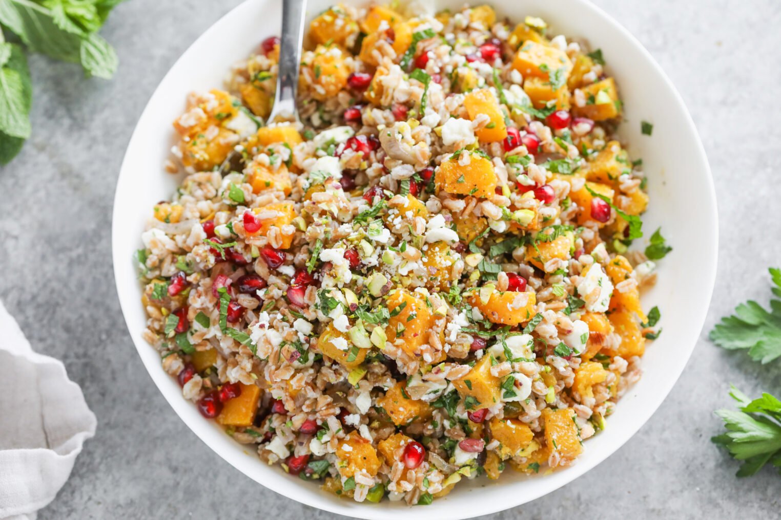 A White Bowl Filled With A Jeweled Farro Salad Featuring Roasted Butternut Squash, Pomegranate Seeds, Crumbled Feta, Chopped Herbs, And A Fork Resting On The Side. The Vibrant Bowl Sits Atop A Gray Surface.