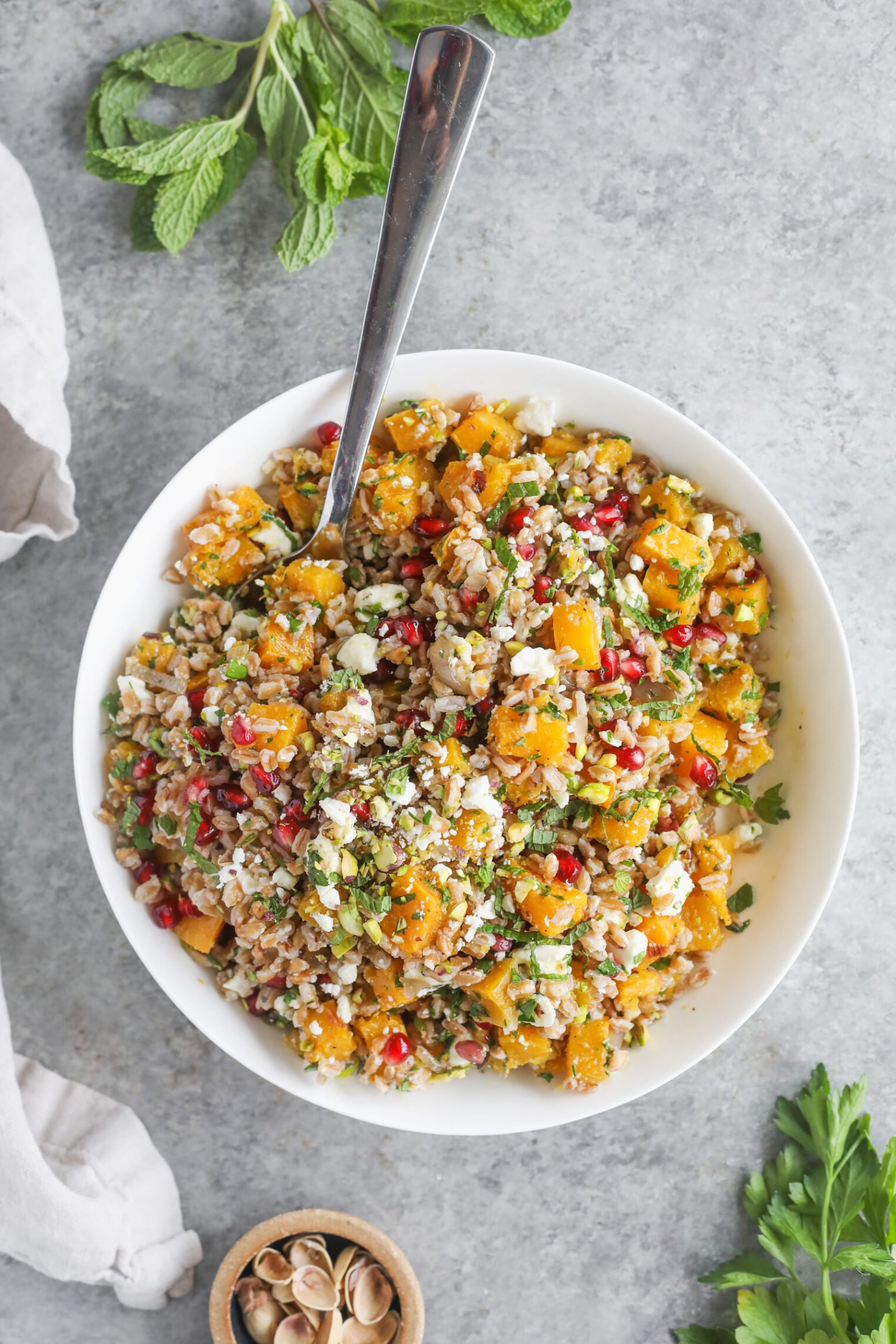 A Bowl Of Colorful Jeweled Farro Salad With Grains, Roasted Butternut Squash, Pomegranate Seeds, Feta Cheese, And Chopped Herbs, With A Spoon In The Bowl. Mint Leaves And Other Ingredients Are Placed Around The Bowl On A Gray Surface.