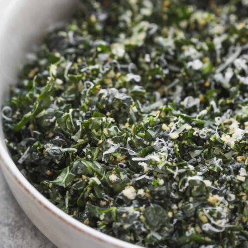 Easy Kale Caesar Salad (6 Ingredients!) 10 A Close-Up Of A Bowl Filled With A Kale Caesar Salad, Topped With Grated Cheese And Small Breadcrumb Pieces. The Salad Is Placed On A Light Gray Surface, Highlighting The Green And Textured Appearance Of The Kale Leaves.