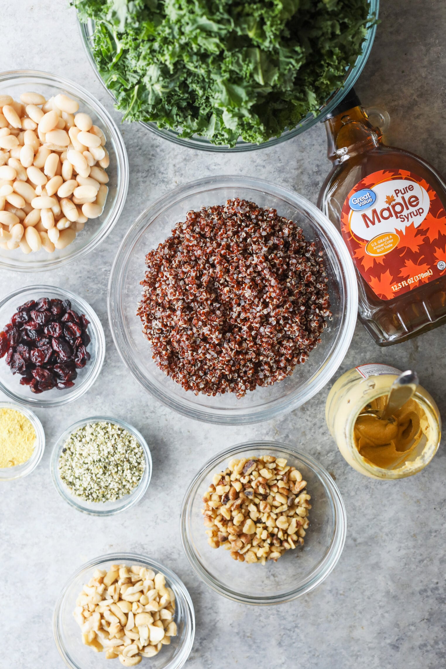 Quinoa Kale Salad 2 Bowls Filled With Kale Salad, Red Quinoa, White Beans, Walnuts, Peanuts, Dried Cranberries, Hemp Seeds, Nutritional Yeast, A Jar Of Mustard, And A Bottle Of Maple Syrup Are Arranged On A Gray Surface.