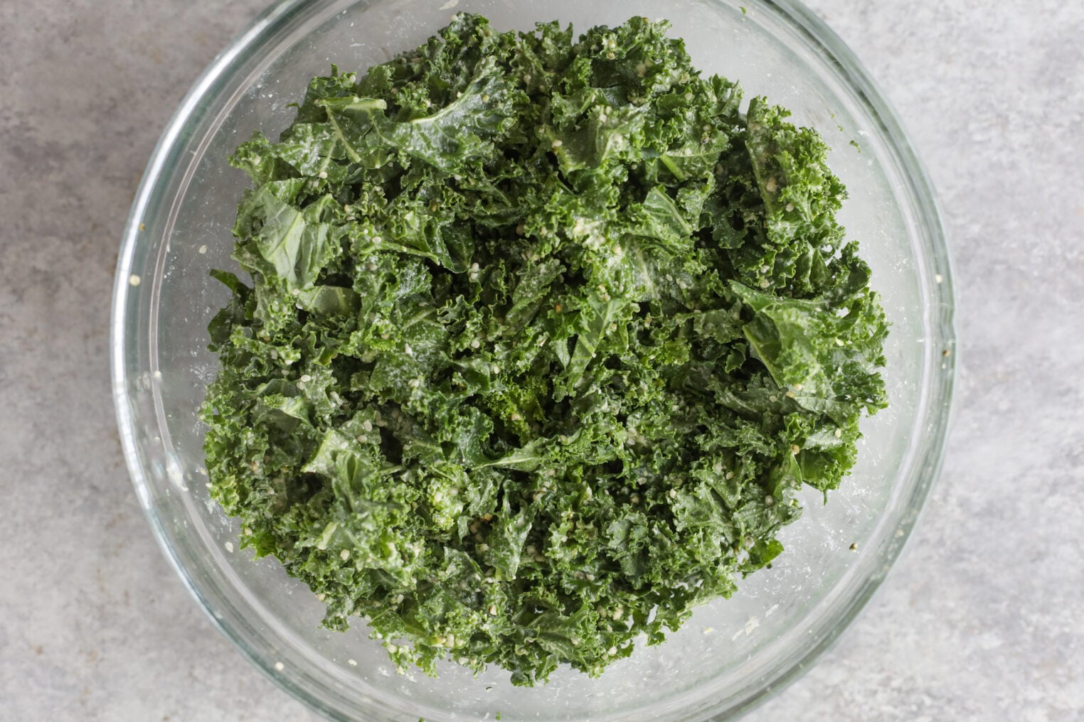 Quinoa Kale Salad 4 A Clear Glass Bowl Filled With Fresh, Chopped Kale Leaves Sits On A Light Gray Surface, Perfect As The Base For A Vibrant Quinoa Kale Salad.