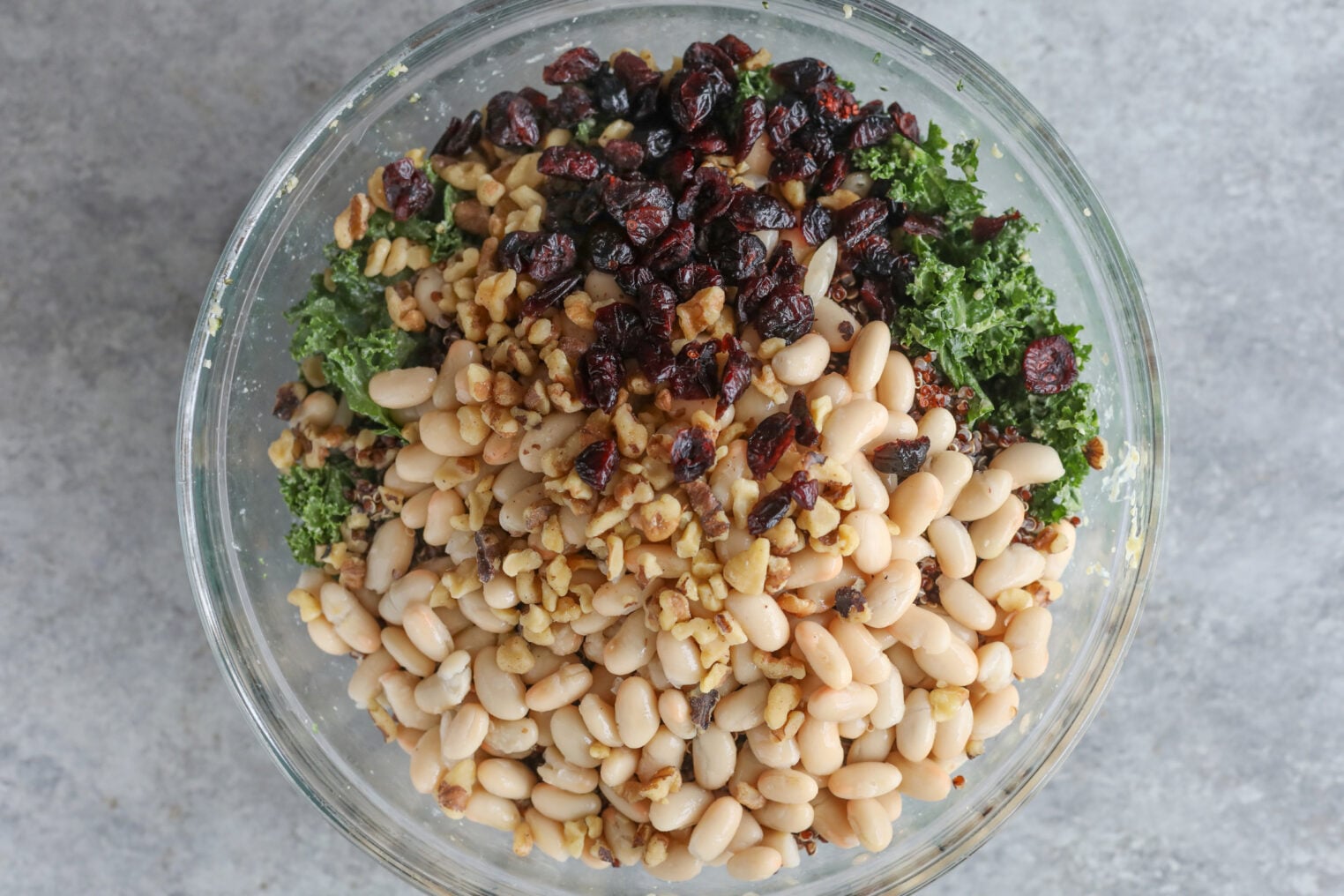 Quinoa Kale Salad 6 A Glass Bowl Filled With White Beans, Chopped Walnuts, Dried Cranberries, And Kale, Layered And Ready To Be Mixed For A Vibrant Quinoa Kale Salad, Sits On A Light Gray Surface.