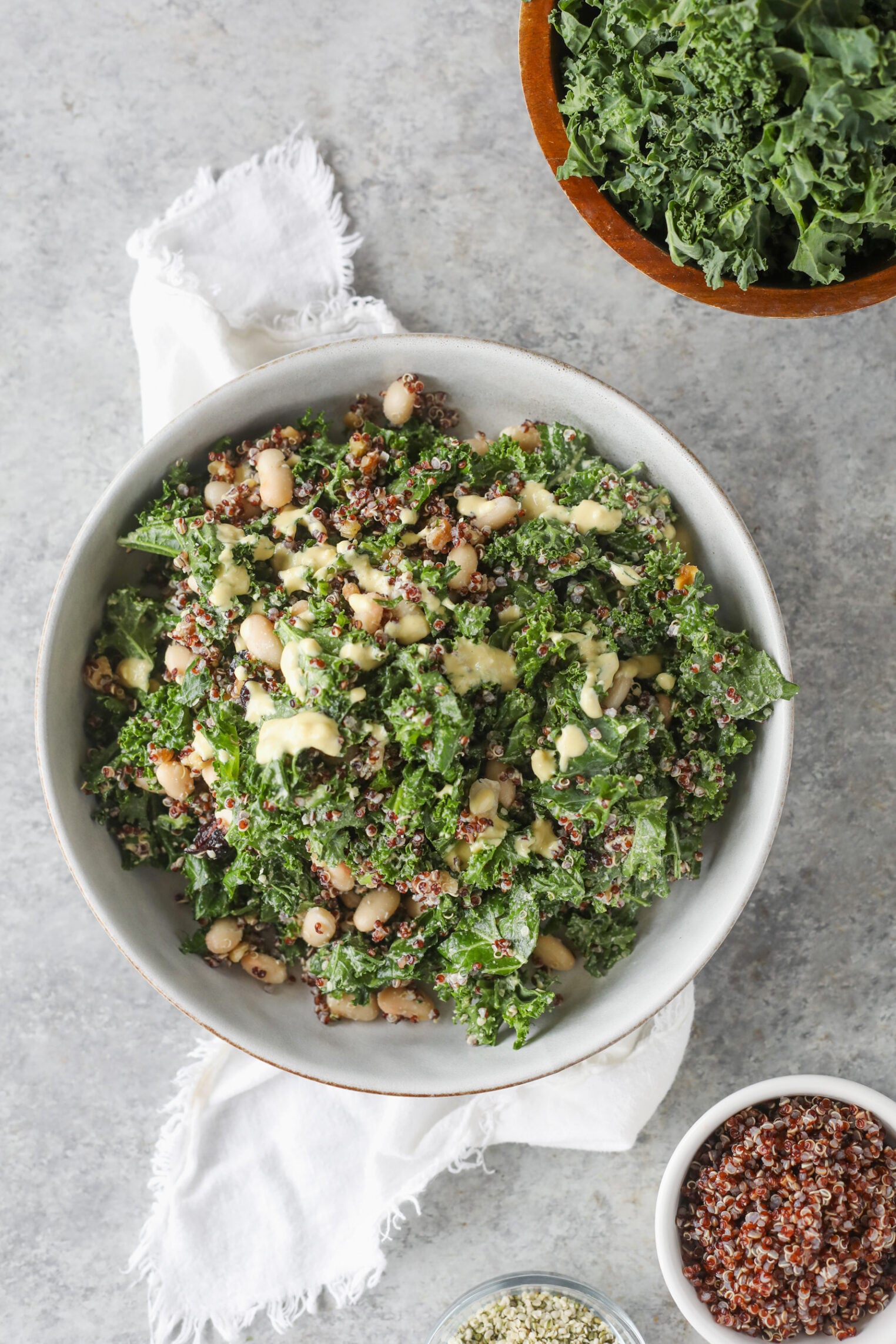 Quinoa Kale Salad 7 A Bowl Of Quinoa Kale Salad With Beans And Creamy Dressing Sits On A Light Surface, Accompanied By Fresh Kale, Small Bowls Of Quinoa And Seeds, And A White Cloth Napkin Nearby.