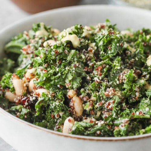 Quinoa Kale Salad 10 A Quinoa Kale Salad Featuring White Beans And Seeds Sits In A Bowl On A Light Surface, With A Wooden Bowl Of Greens And A Small Glass Dish In The Background.