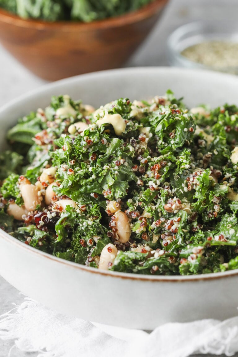 A Quinoa Kale Salad Featuring White Beans And Seeds Sits In A Bowl On A Light Surface, With A Wooden Bowl Of Greens And A Small Glass Dish In The Background.