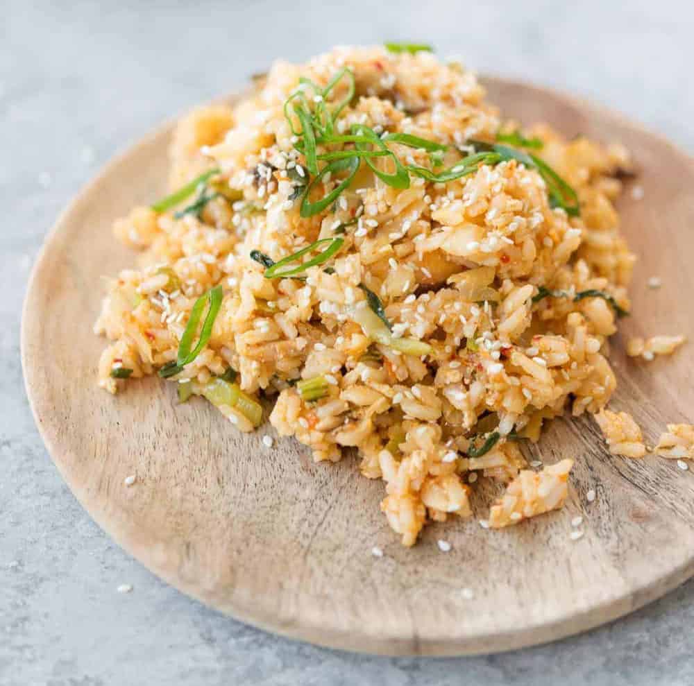 A Wooden Plate Filled With Fried Rice, A Staple In Healthy Vegetarian Meals, Is Garnished With Chopped Green Onions And Sprinkled With Sesame Seeds. The Rice Incorporates Small Pieces Of Vegetables And Boasts A Slightly Golden, Seasoned Appearance.