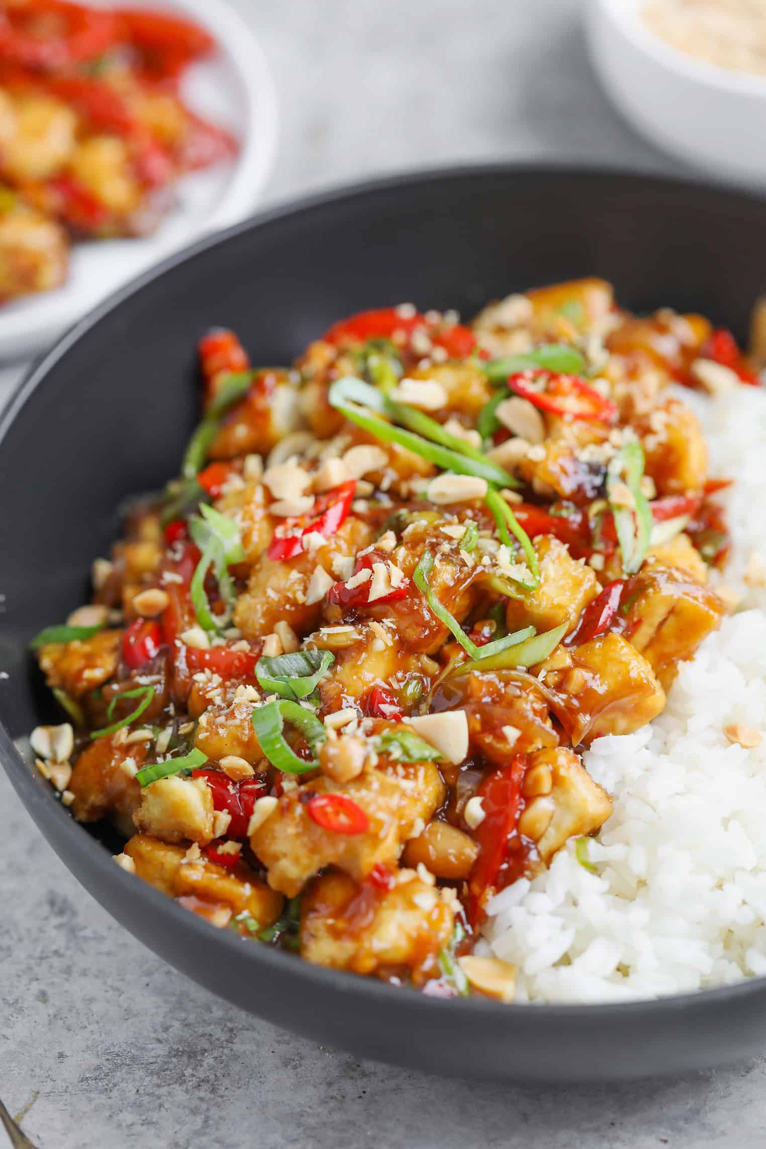 A Black Bowl Filled With White Rice And A Colorful Vegan Kung Pao Stir-Fry Featuring Tofu, Red Bell Peppers, Chopped Green Onions, And Crushed Peanuts, All Coated In A Glossy Sauce.