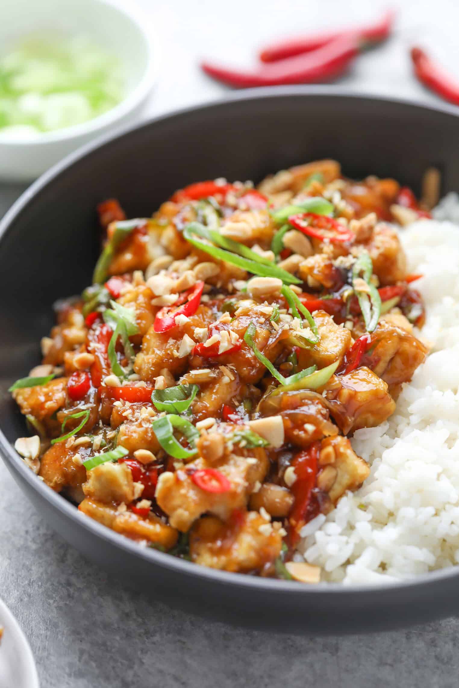 Kung Pao Tofu 3 A Bowl Of White Rice Topped With Kung Pao Tofu, Chopped Peanuts, Sliced Red And Green Chilies, And Green Onions, All Garnished With A Savory Sauce.