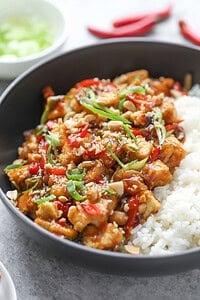A Bowl Of White Rice Topped With Kung Pao Tofu, Chopped Peanuts, Sliced Red And Green Chilies, And Green Onions, All Garnished With A Savory Sauce.