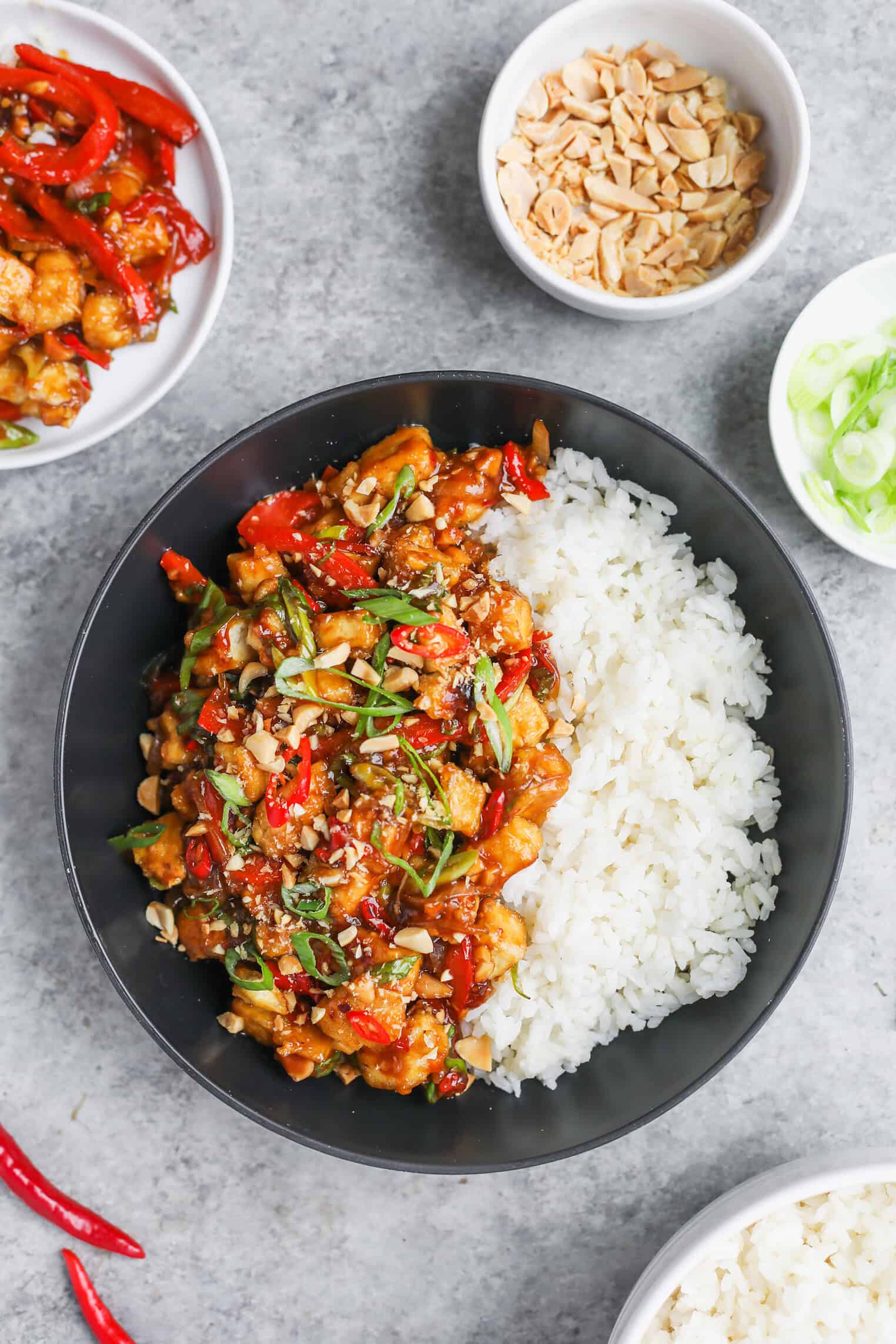 Kung Pao Tofu 12 A Black Bowl Filled With White Rice And A Colorful Stir-Fry Of Chicken, Red Bell Peppers, Green Onions, And Crushed Peanuts—Perfect For Pairing With Kung Pao Tofu. Small Dishes Of Peanuts, Green Onions, And Extra Stir-Fry Sit On A Gray Surface.