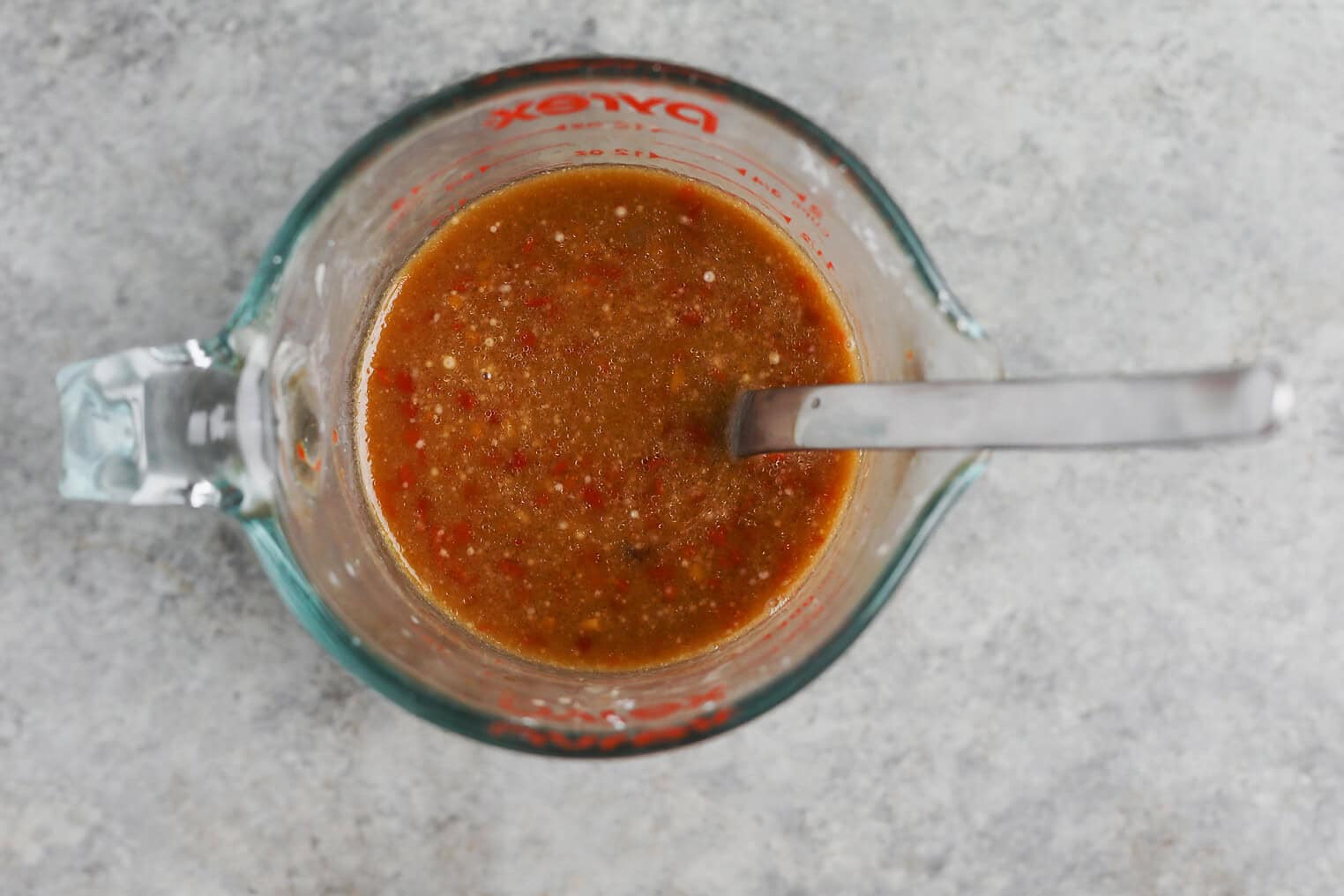 Kung Pao Tofu 5 A Glass Measuring Cup Filled With A Brownish-Red Kung Pao Tofu Sauce Mixture Containing Visible Spices And Small Bits, With A Metal Spoon Resting Inside, On A Gray Textured Surface.