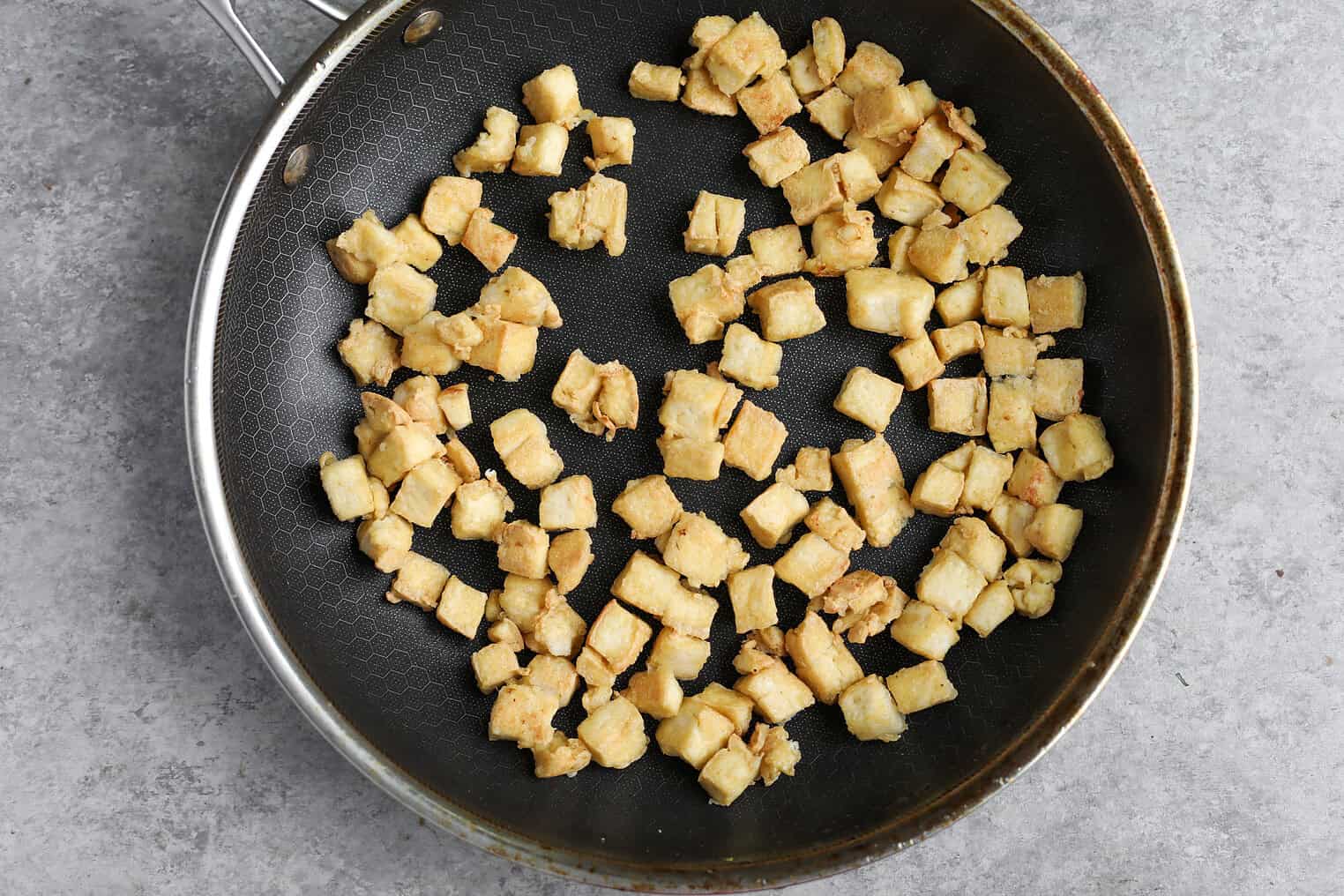 Kung Pao Tofu 7 Cubed Pieces Of Tofu, Perfect For Kung Pao Tofu, Are Spread Out And Lightly Browned In A Nonstick Frying Pan, Resting On A Grey Textured Surface.