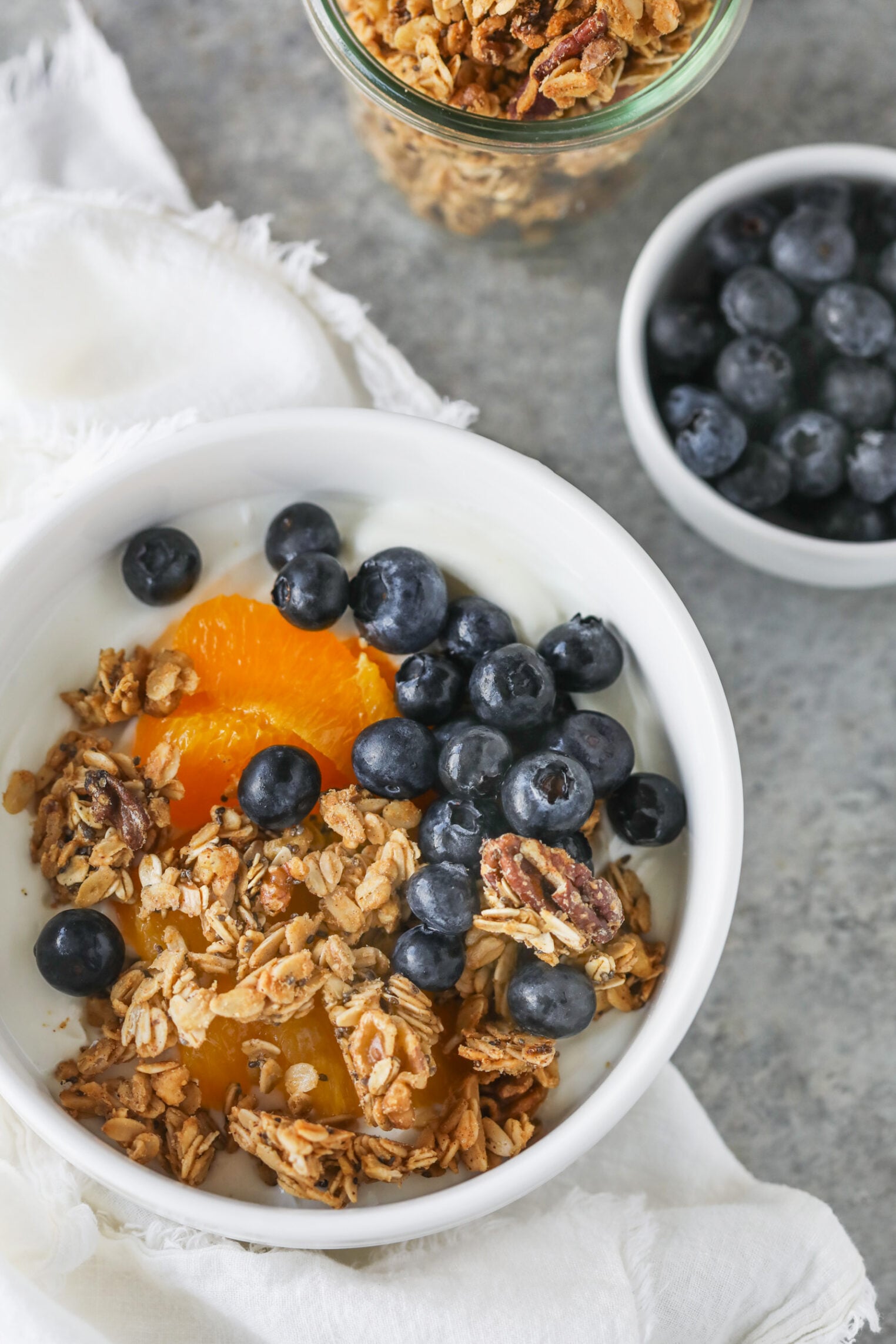 A White Bowl With Yogurt Topped With Low Sugar Granola, Fresh Blueberries, And Orange Slices. Nearby, There Is A Small Bowl Of Blueberries And A Jar Of Low Sugar Granola On A Gray Surface With A White Napkin.
