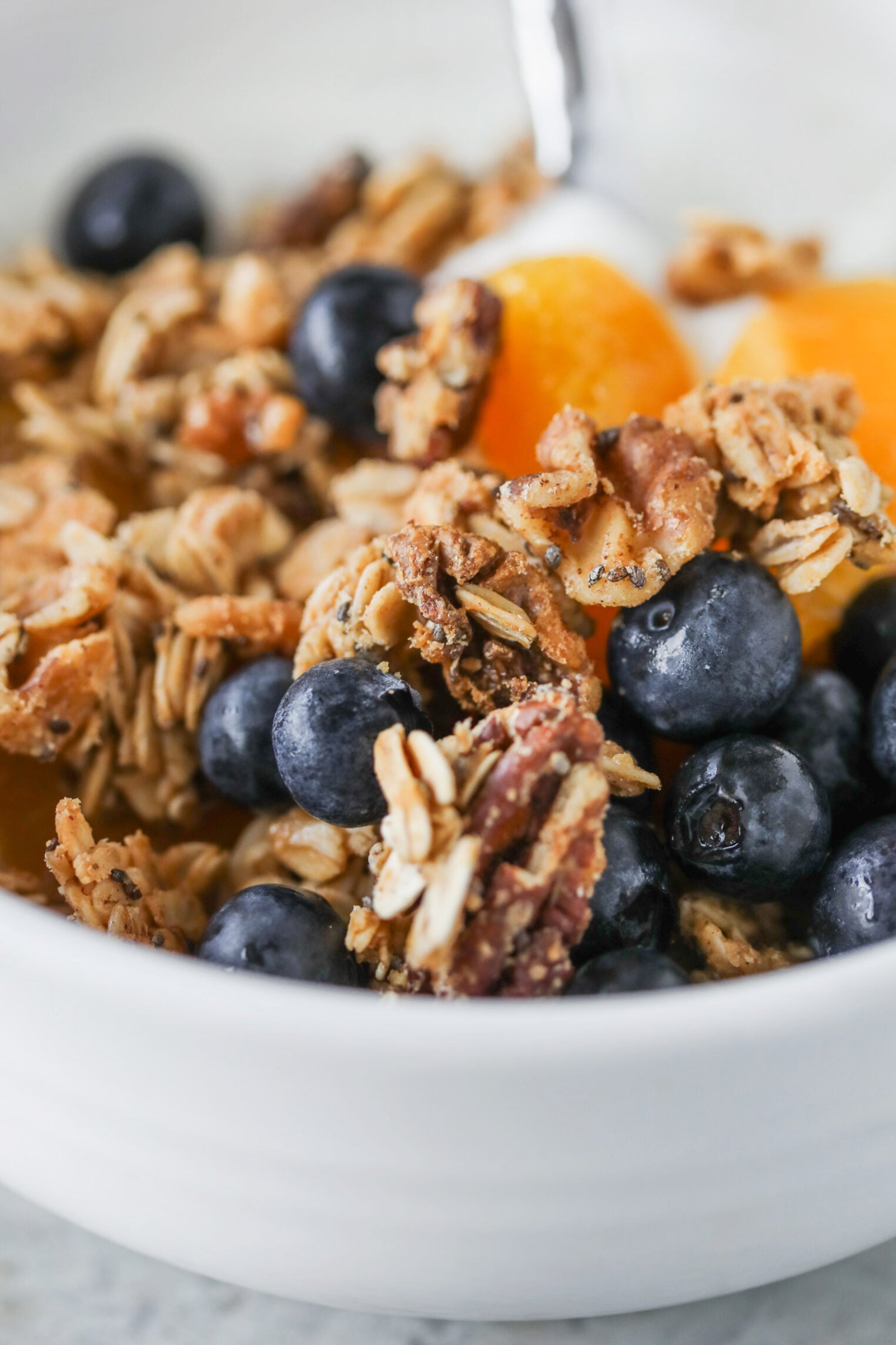 A Close-Up Of A White Bowl Filled With Low Sugar Granola Clusters, Fresh Blueberries, And Orange Fruit Segments, With A Spoon Partially Visible In The Background.