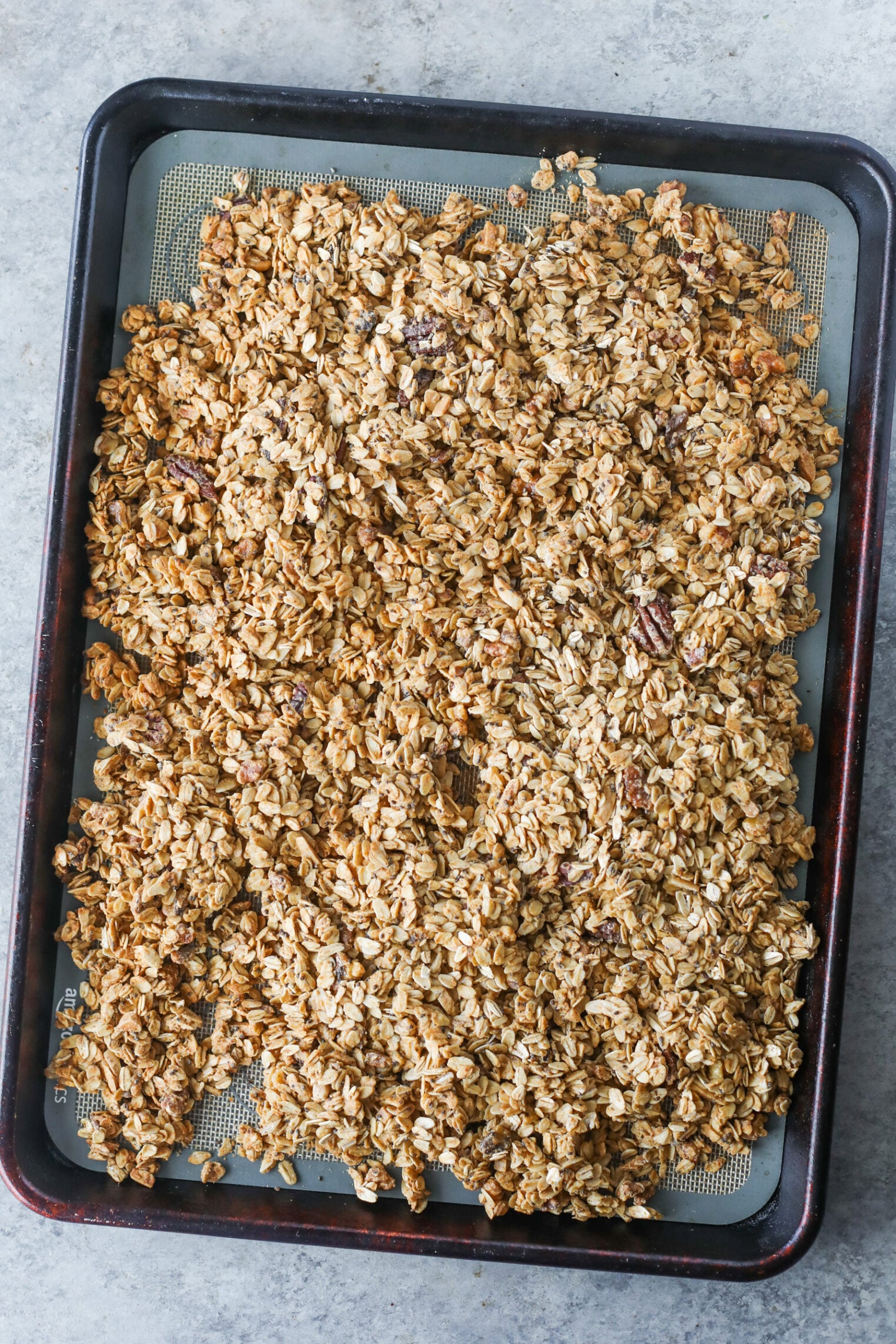 A Baking Sheet Covered With A Layer Of Golden-Brown Low Sugar Granola, Spread Evenly Over A Silicone Baking Mat, Resting On A Gray Countertop.