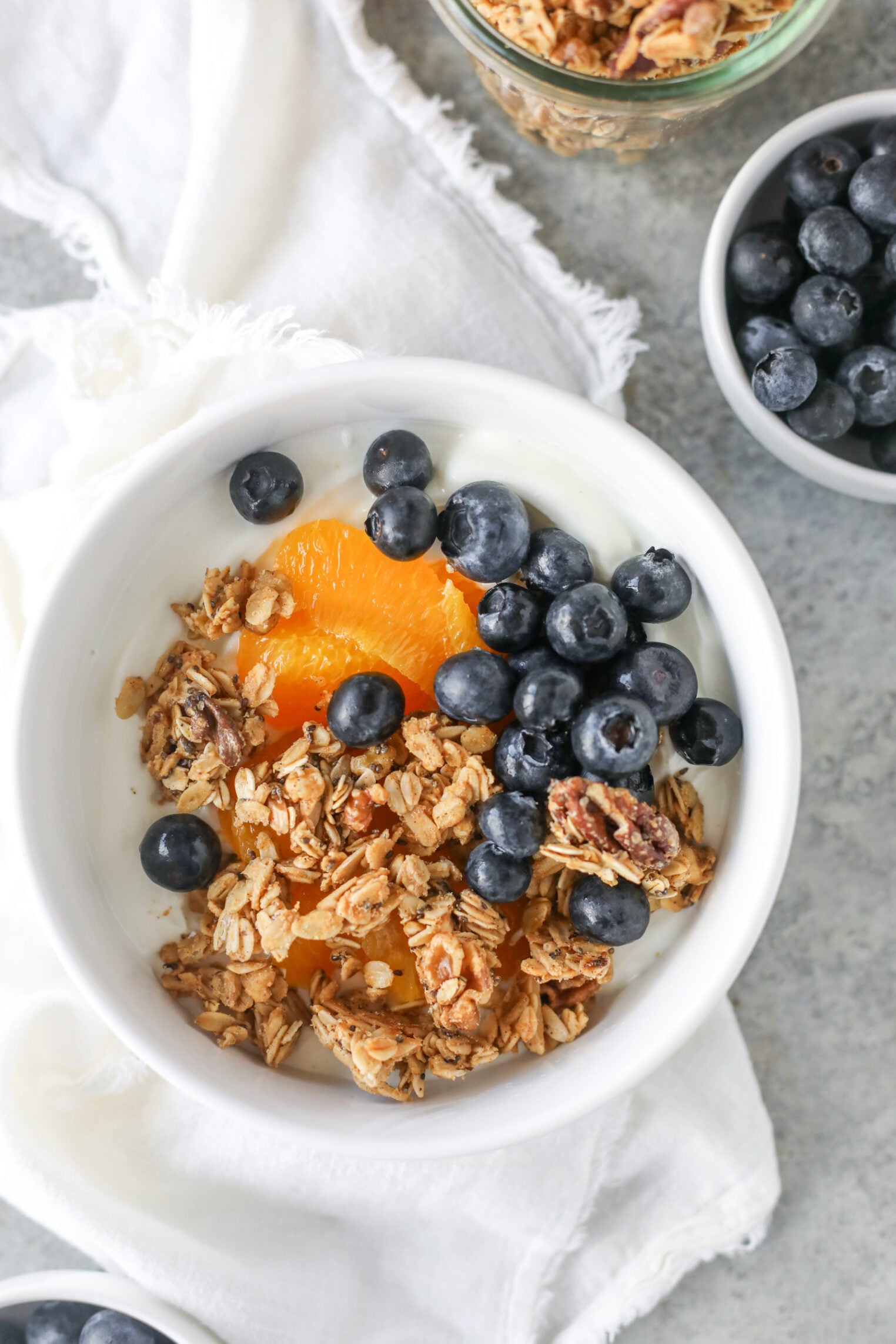 A White Bowl Filled With Yogurt, Low Sugar Granola, Peach Slices, And Fresh Blueberries, Placed On A White Cloth With Extra Bowls Of Granola And Blueberries Nearby.