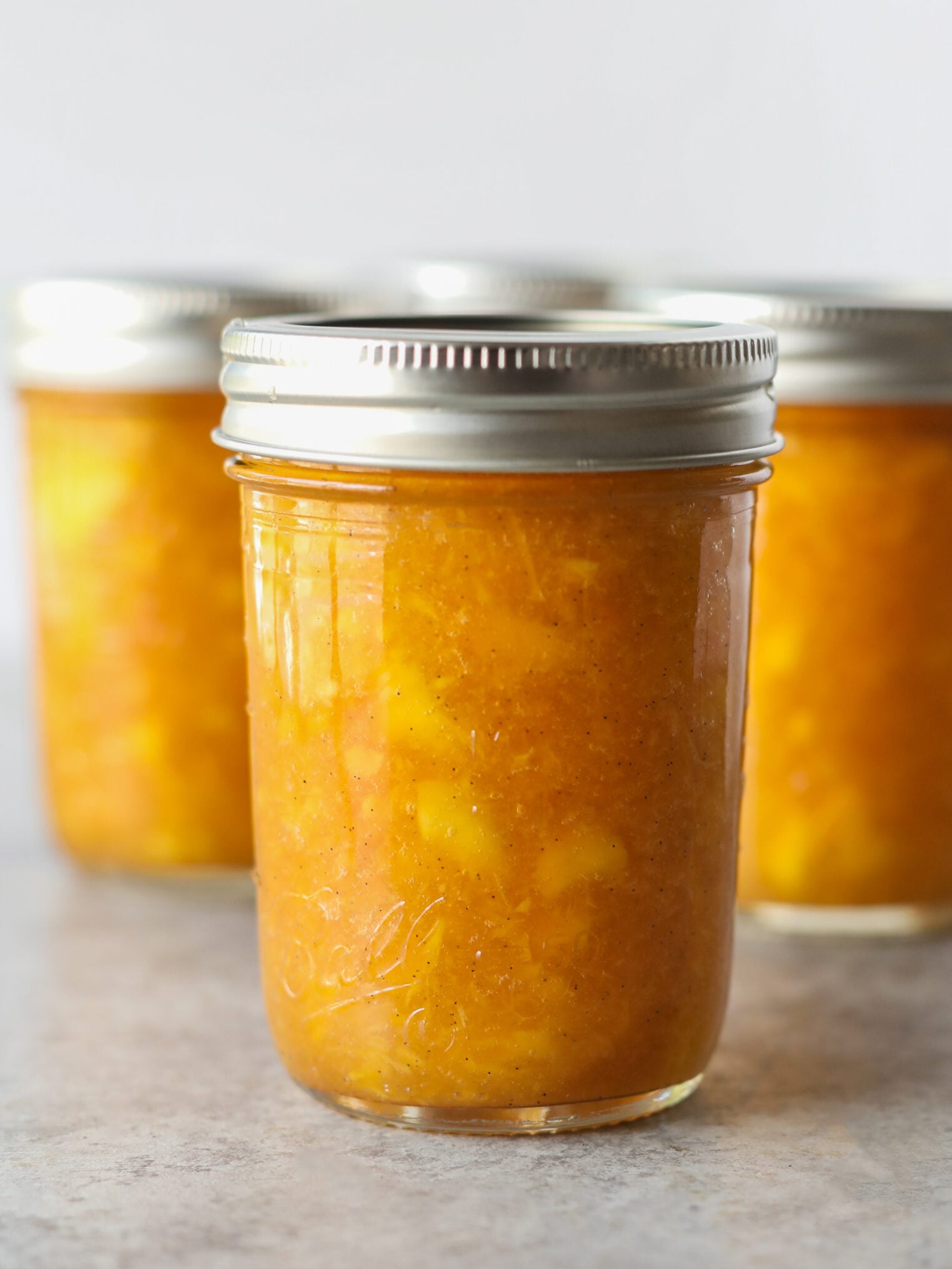 A Glass Jar Filled With Vibrant Low Sugar Peach Jam, Sealed With A Silver Metal Lid, Sits In Front Of Two Similar Jars On A Light Surface.