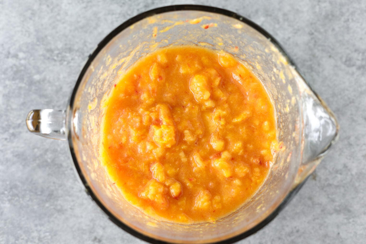 A Glass Bowl Filled With A Chunky, Orange-Yellow Mashed Mixture—Reminiscent Of Low Sugar Peach Jam Or Possibly Cooked Squash Or Sweet Potatoes—Sits On A Gray, Textured Surface.