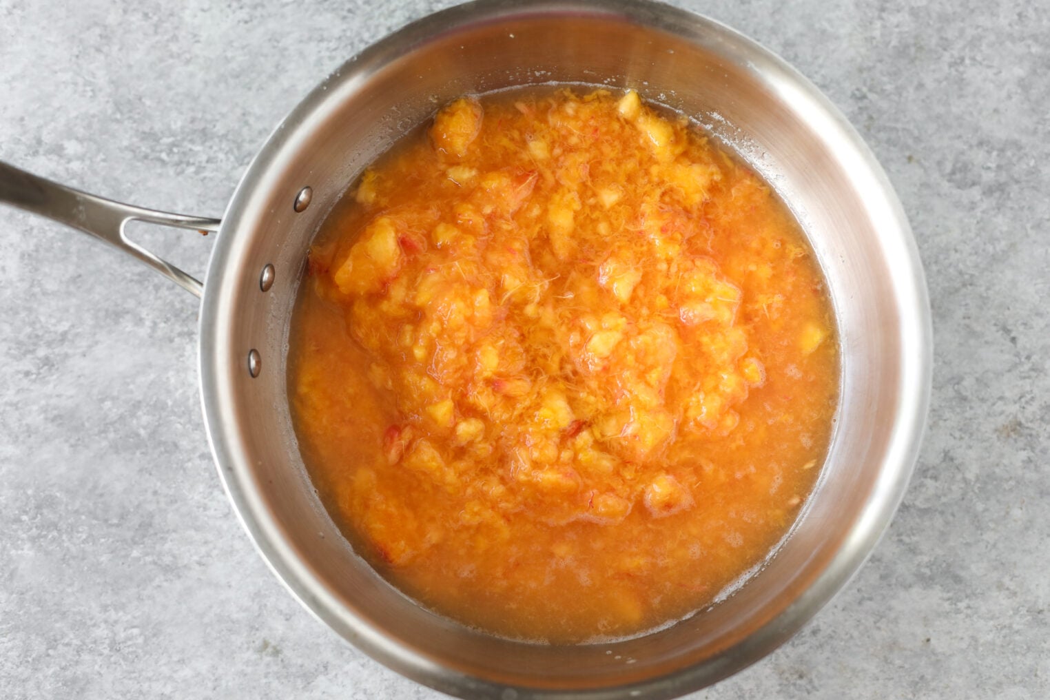 A Stainless Steel Saucepan Filled With Chunky Orange And Yellow Fruit Mixture, Likely Low Sugar Peach Jam Being Cooked Down, Sits On A Gray Countertop.