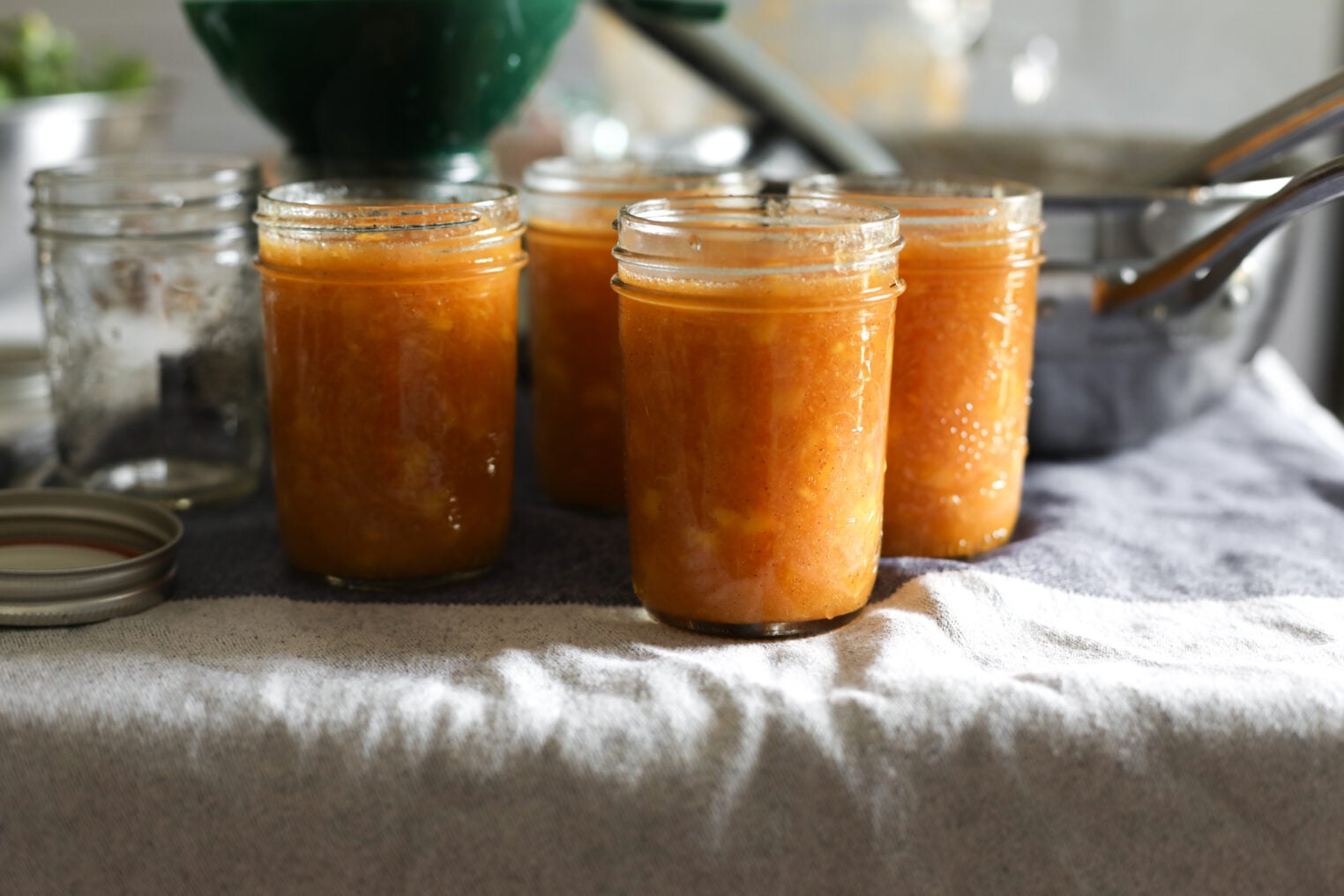Four Glass Jars Filled With Low Sugar Peach Jam Are Set On A Towel, With An Empty Jar, A Lid, And Kitchen Utensils Visible In The Background.