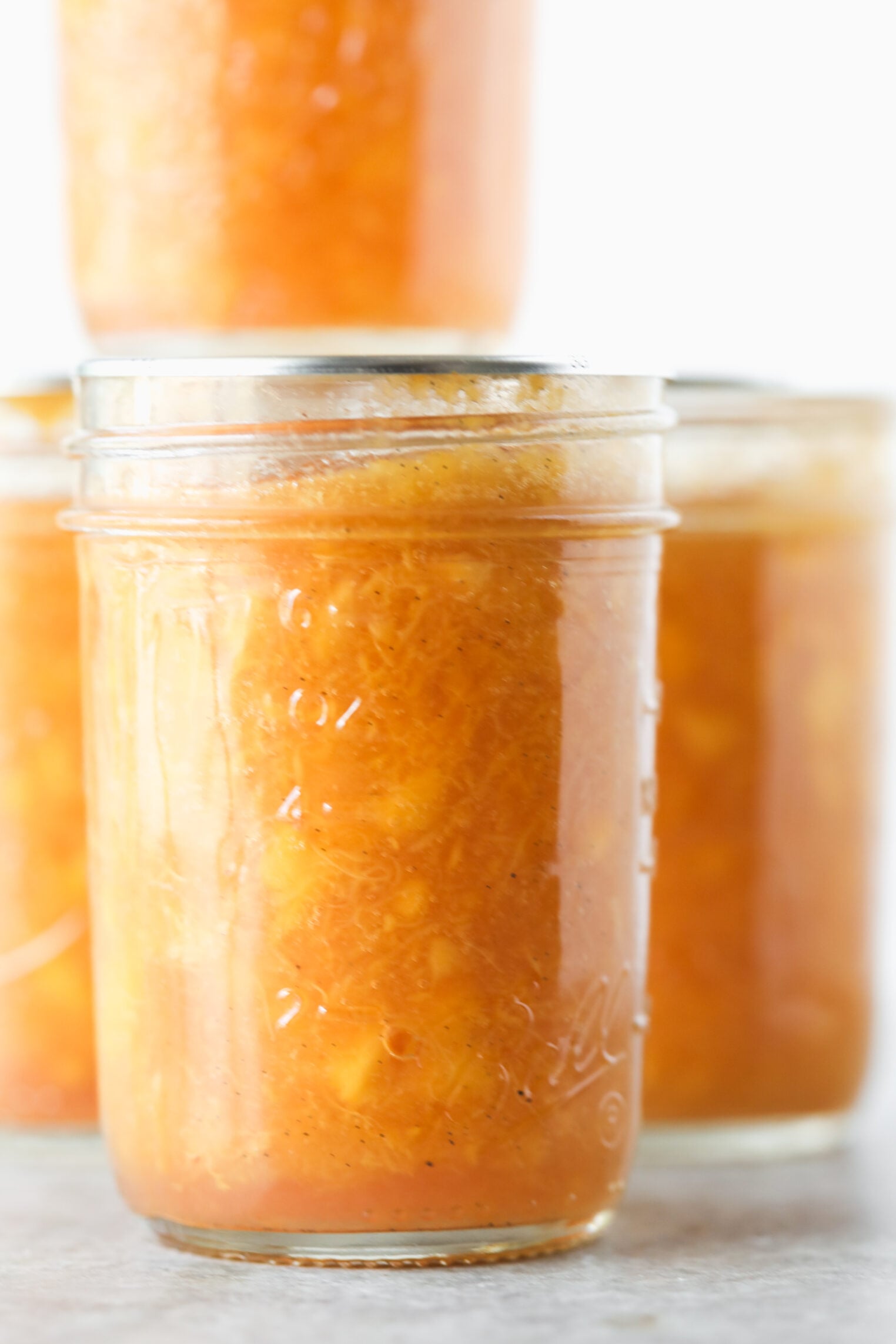 Three Glass Jars Filled With Vibrant, Orange-Colored Low Sugar Peach Jam Or Preserves, With One Jar Stacked On Top In The Background, Against A White Backdrop.