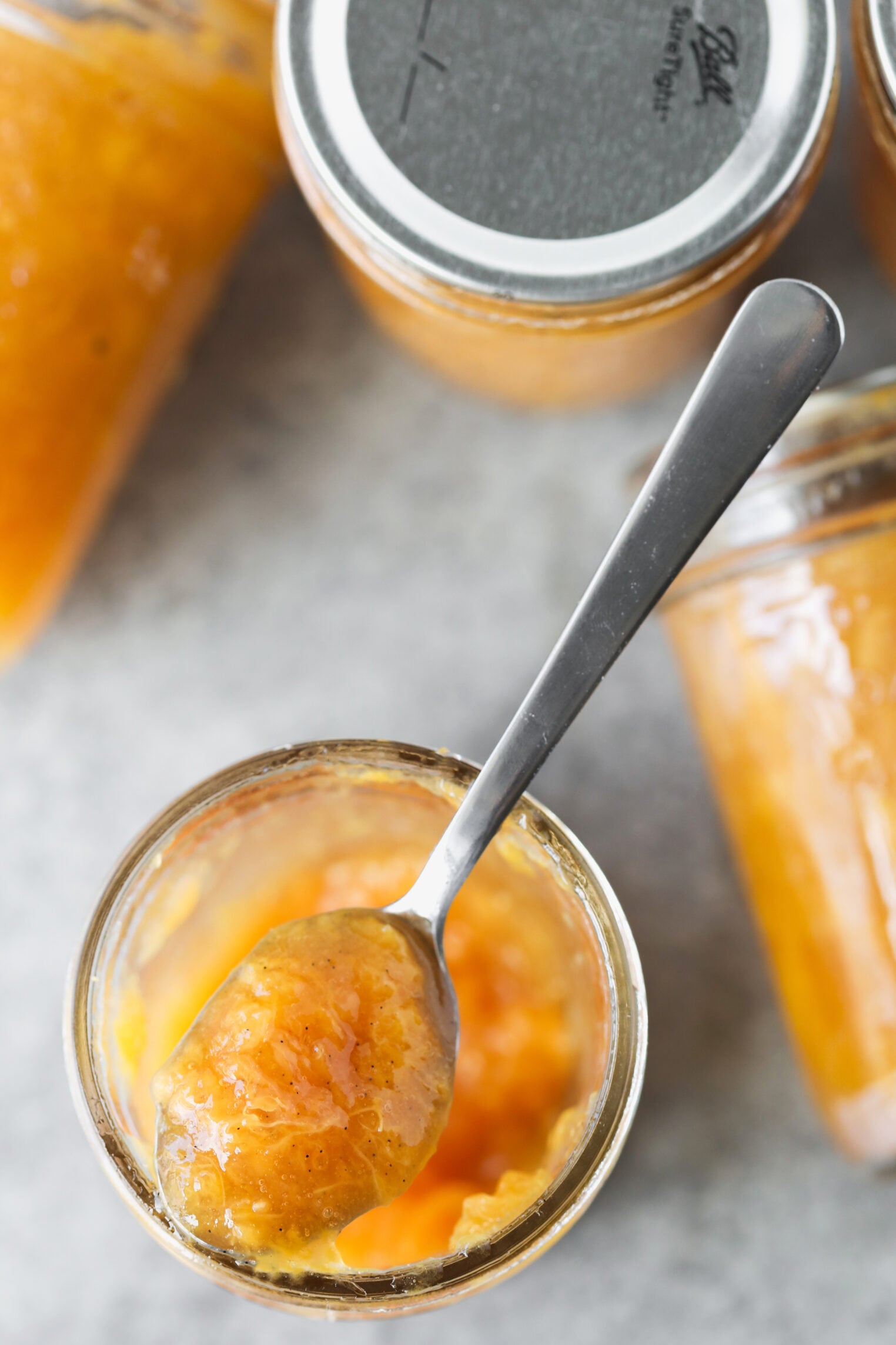 A Spoonful Of Orange-Colored Low Sugar Peach Jam Is Held Over An Open Glass Jar, Surrounded By Other Jars Filled With The Same Jam On A Gray Surface.