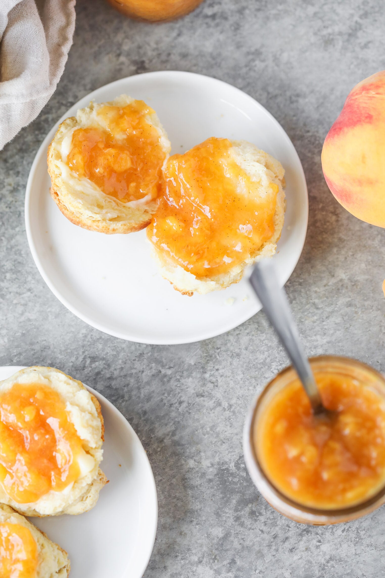 Two Biscuit Halves Topped With Low Sugar Peach Jam Sit On A White Plate. A Jar Of Jam With A Spoon Is Nearby, Along With Another Plate Holding More Biscuits And A Fresh Peach On A Gray Surface.