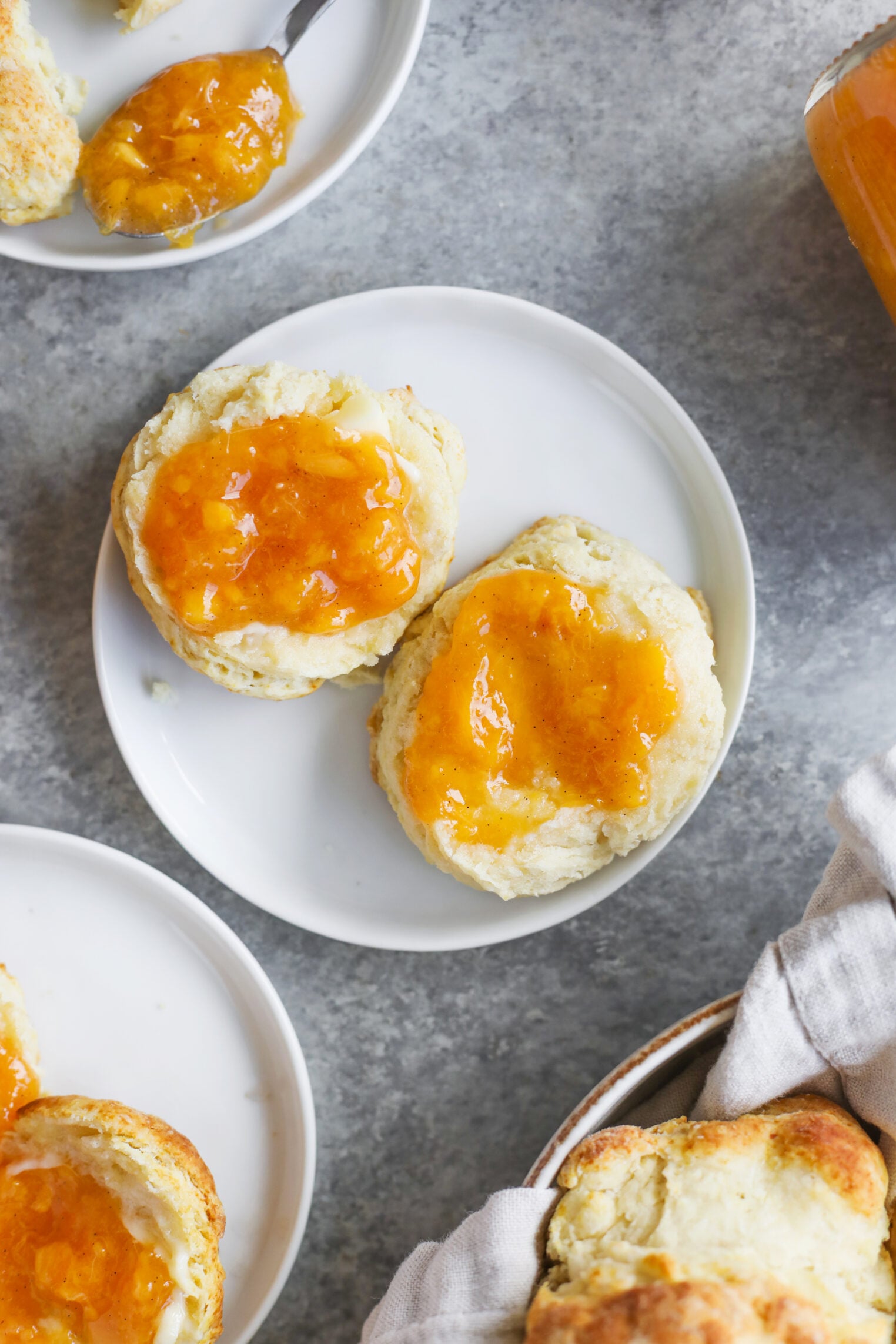 Two Biscuits Split Open On A White Plate, Each Topped With Golden Orange Low Sugar Peach Jam. More Biscuits And A Jar Of Jam Are Visible Nearby On A Gray Surface.