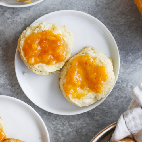 Two Biscuits Split Open On A White Plate, Each Topped With Golden Orange Low Sugar Peach Jam. More Biscuits And A Jar Of Jam Are Visible Nearby On A Gray Surface.