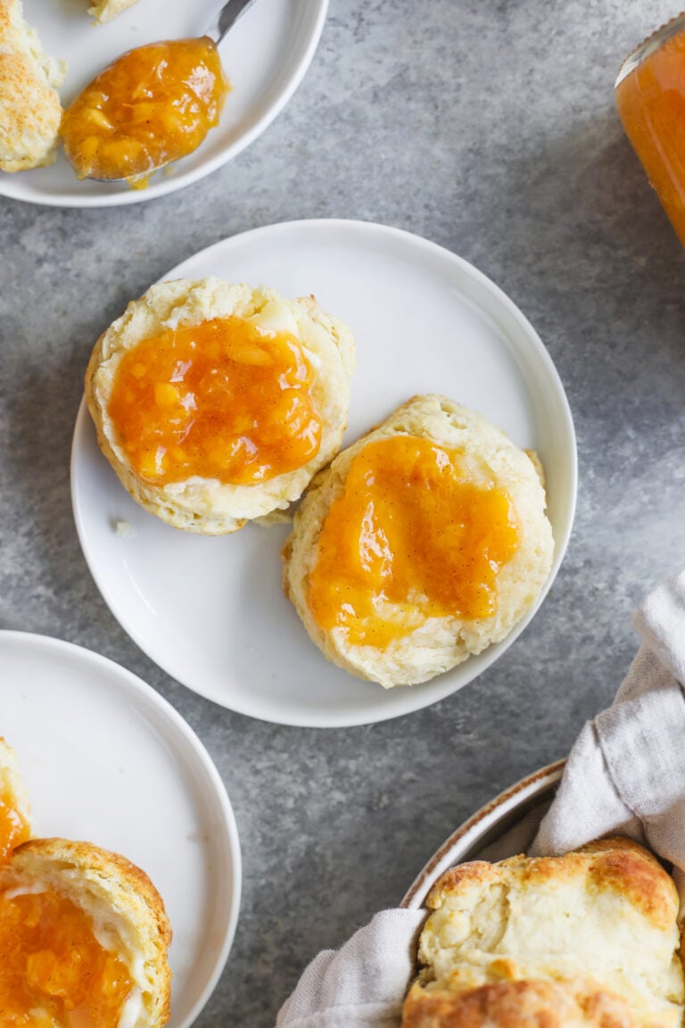 Two Biscuits Split Open On A White Plate, Each Topped With Golden Orange Low Sugar Peach Jam. More Biscuits And A Jar Of Jam Are Visible Nearby On A Gray Surface.